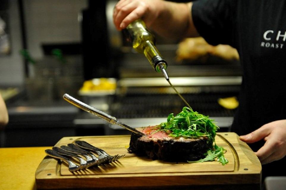 A hand drizzles oil over a juicy, grilled steak topped with fresh arugula on a wooden board. Forks lie beside, suggesting anticipation and savoriness.