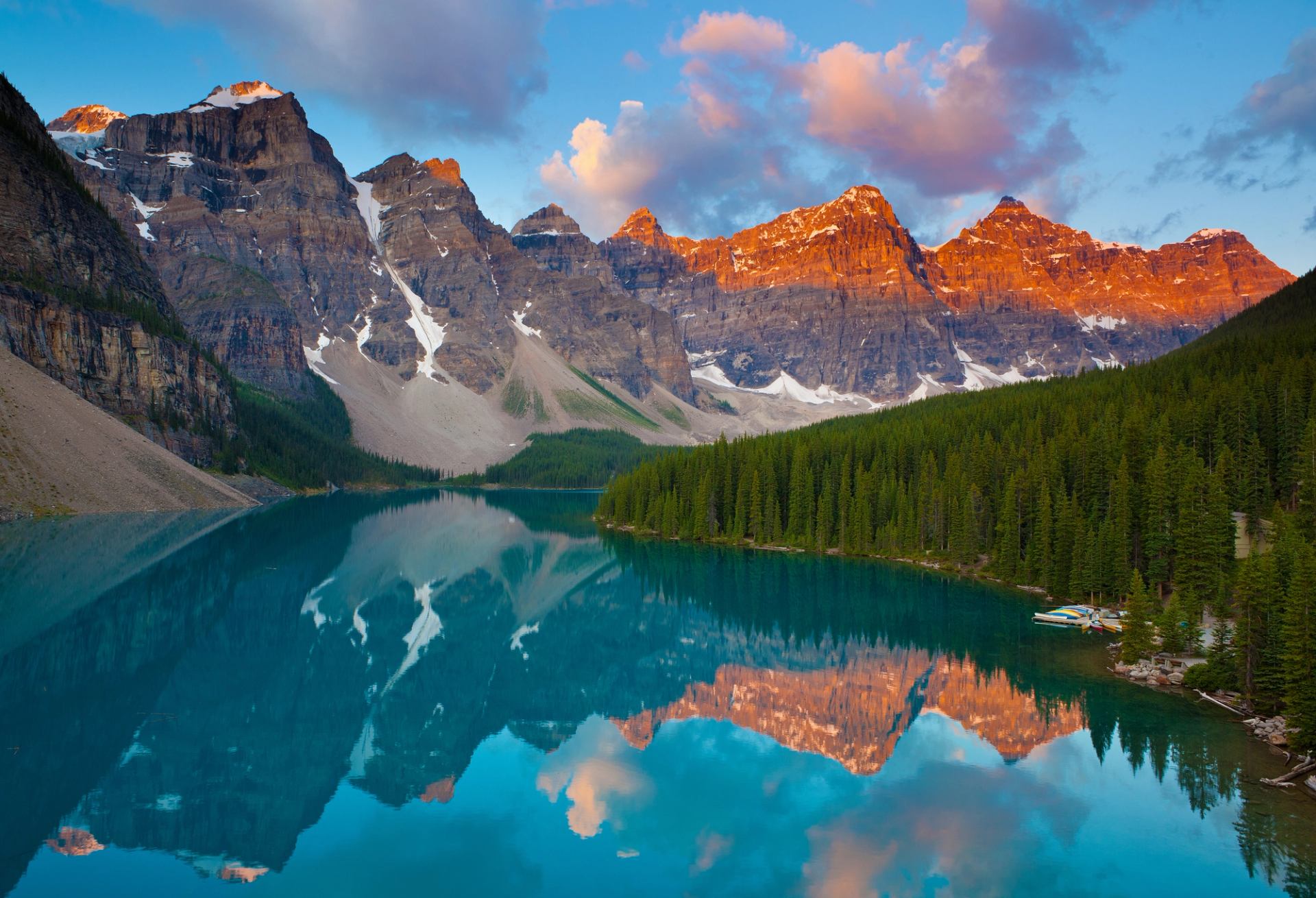 Rocky mountain peaks with snow reflect in a calm turquoise lake beside dense forest.