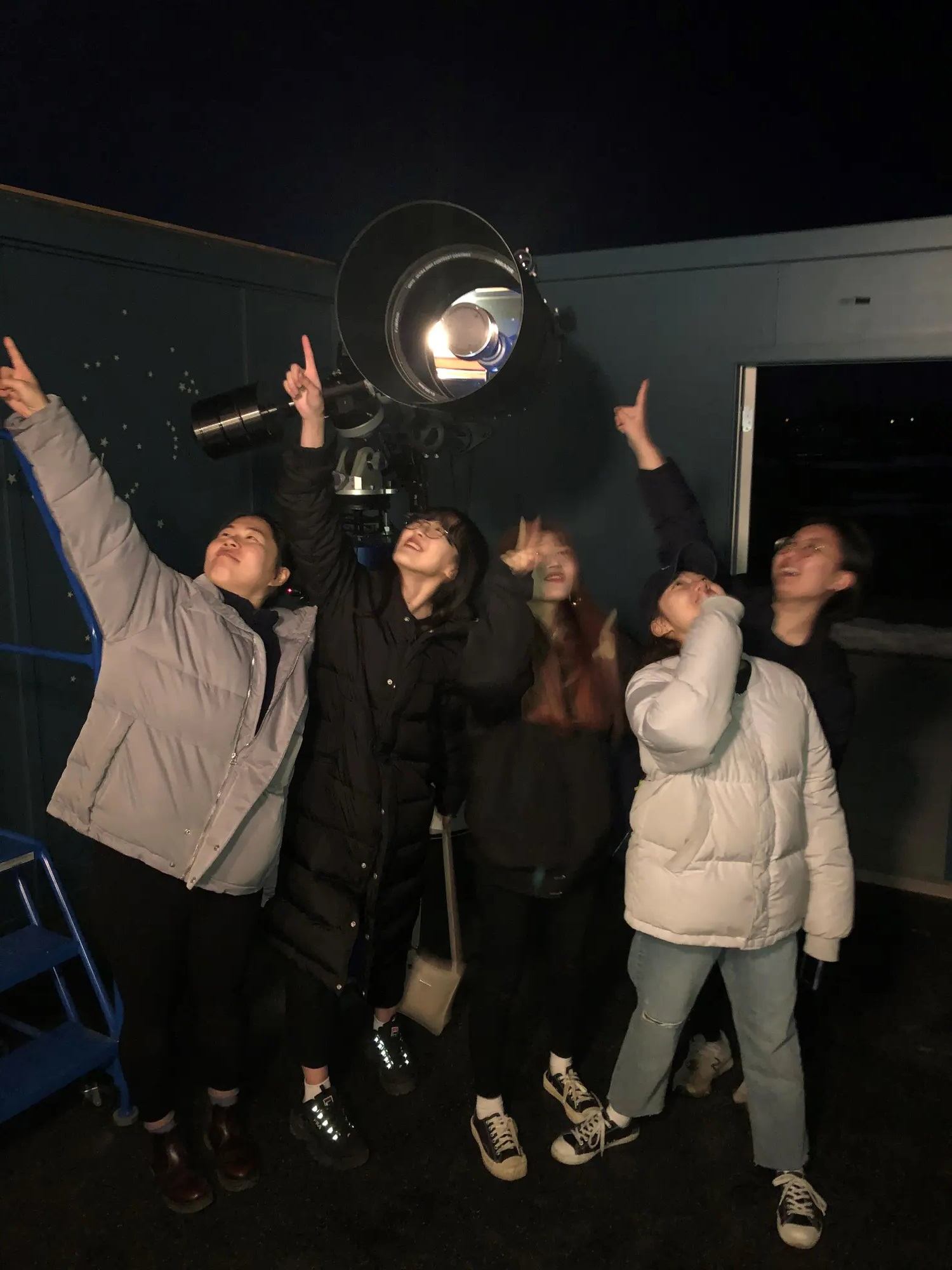 Group stargazing at night with telescope in background.