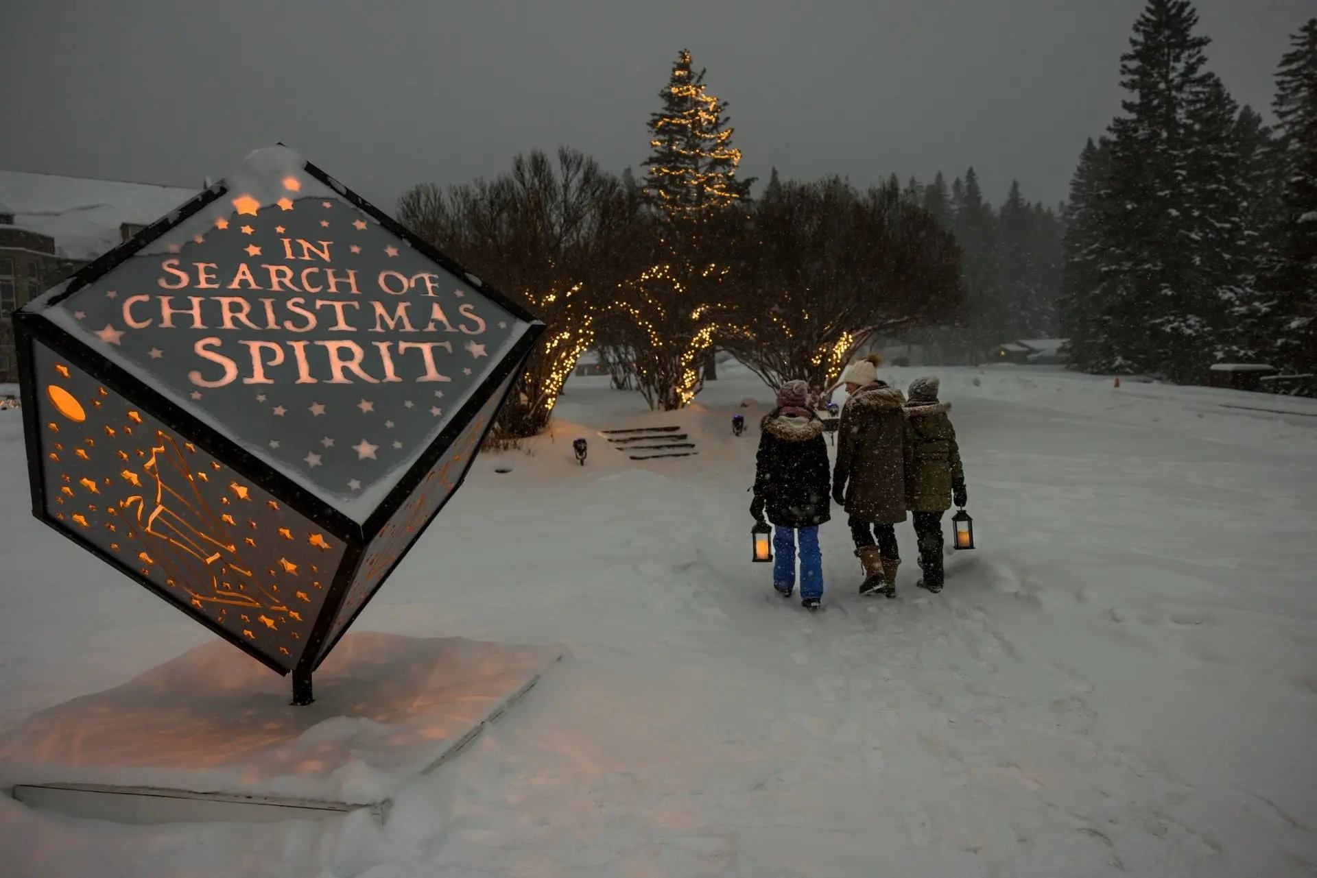 Three people with lanterns walk toward a lit Christmas tree past a glowing cube in snowy twilight.