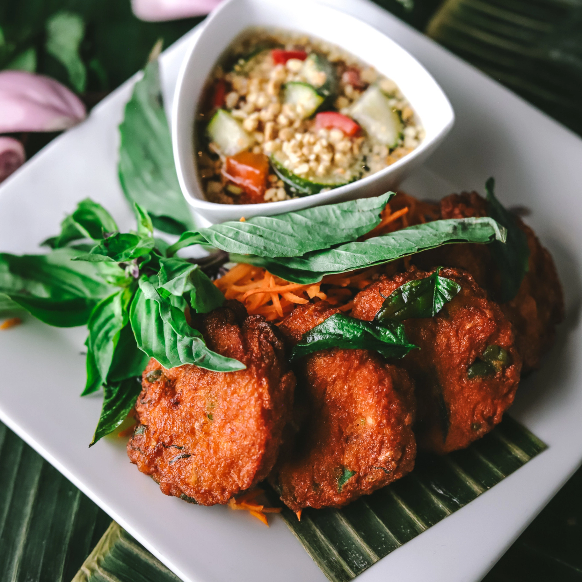 Plate of fried fish cakes with basil leaves and shredded carrots beside dipping sauce.