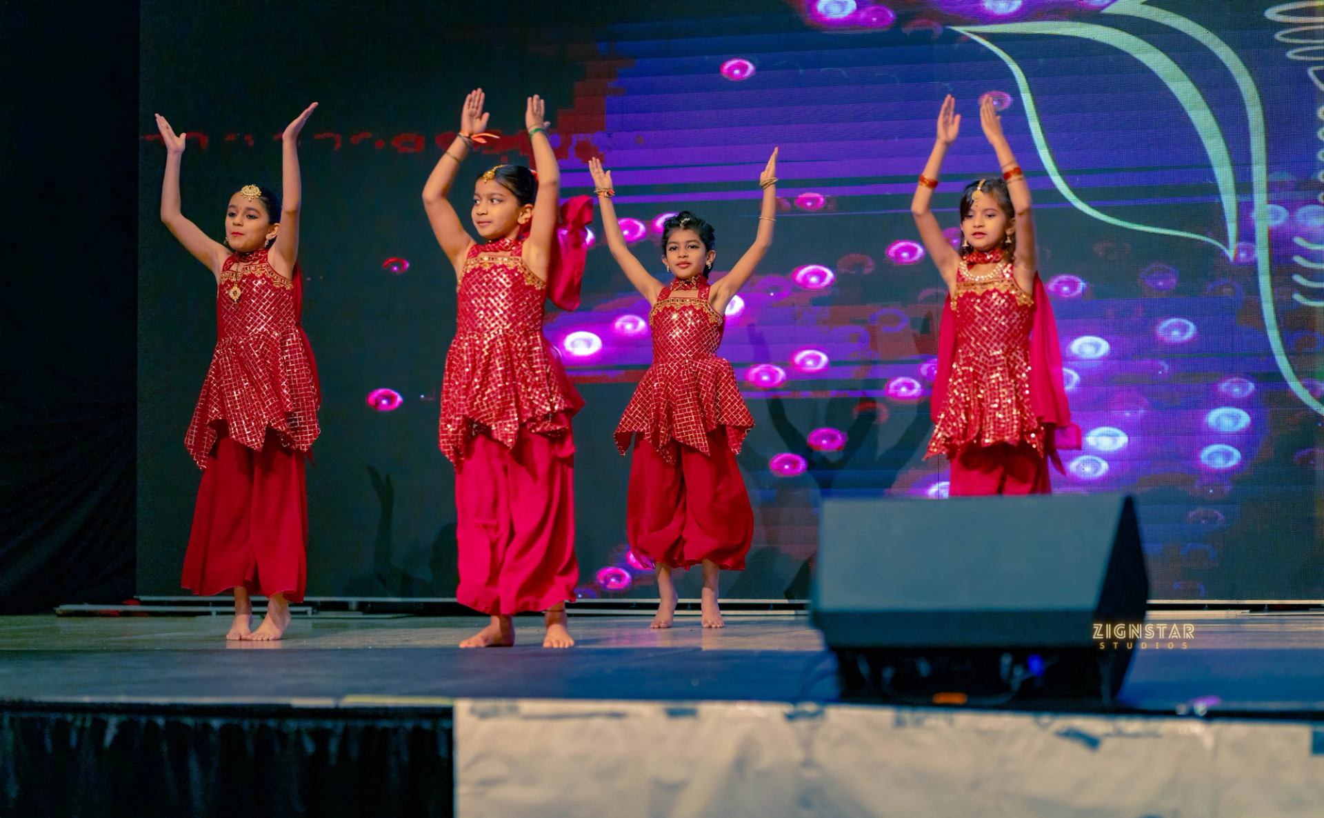 Children in red costumes dance with raised arms on a brightly lit stage.