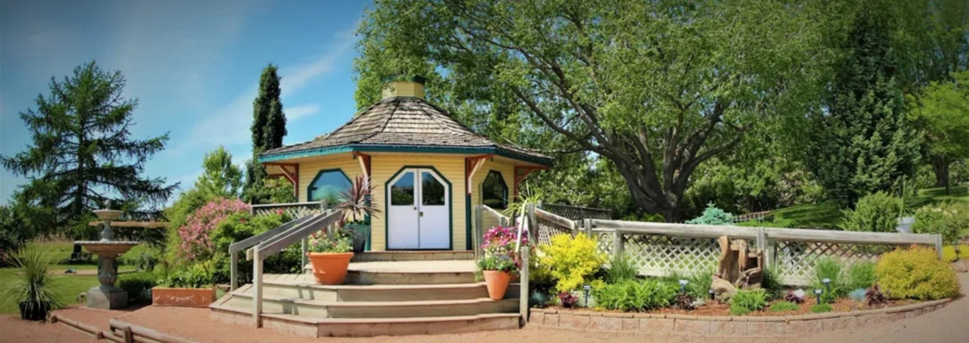Octagonal garden gazebo with ramp, surrounded by colourful flowers, shrubs and large trees.