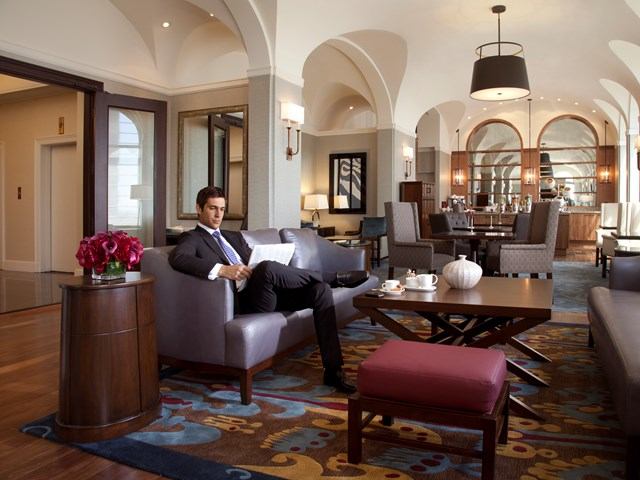 A man in a suit sits on a modern sofa in an elegant, arch-ceilinged lounge. The room features stylish furniture, soft lighting, and vibrant decor.