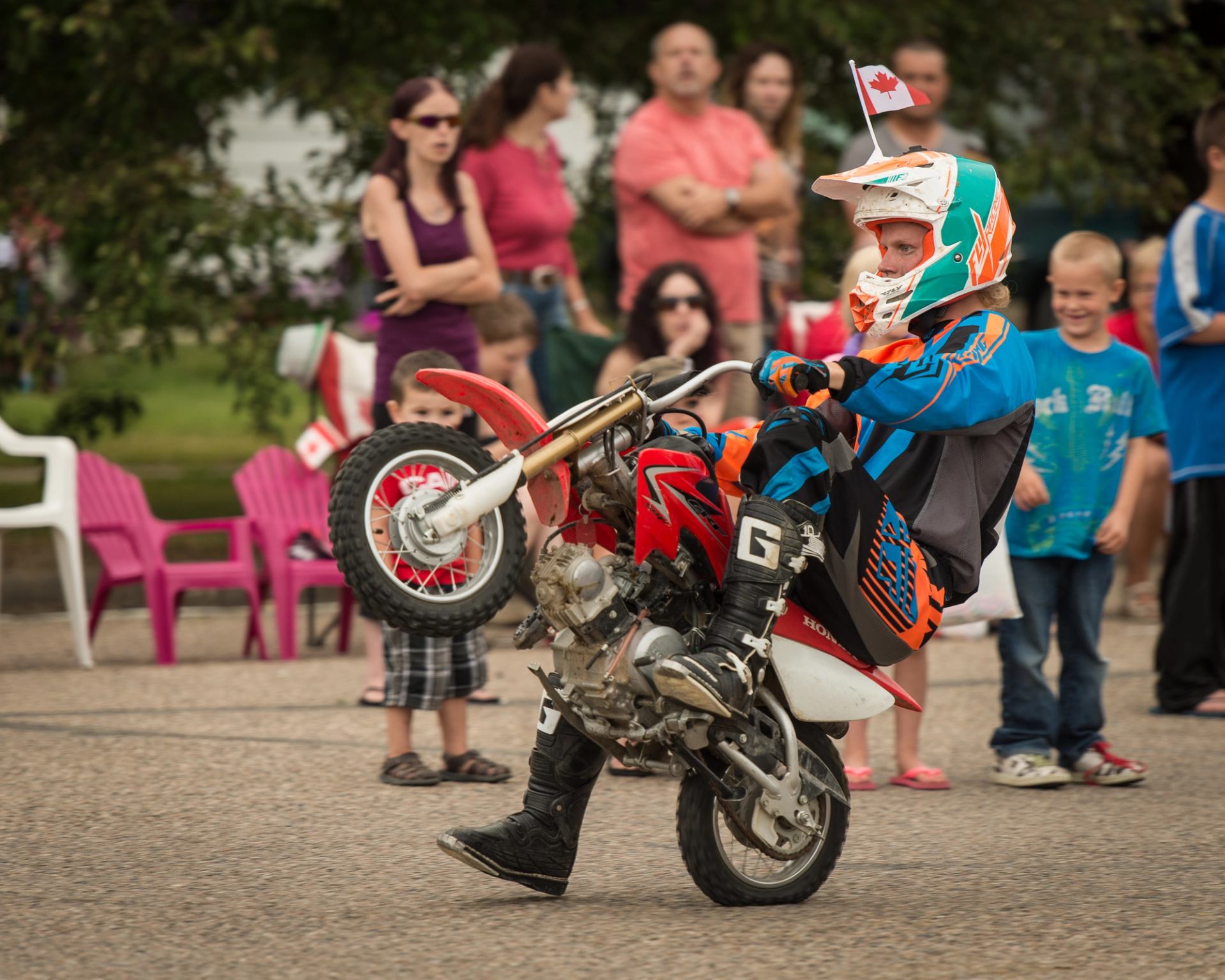 Motocross rider performs wheelie on small dirt bike before crowd with Canadian flags.