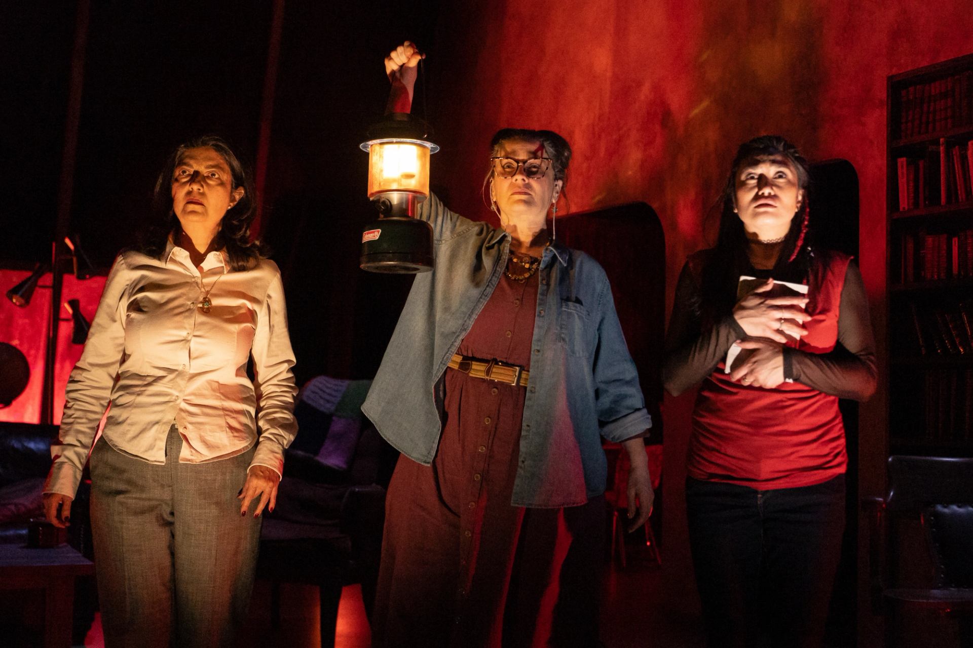 Three people on a dim stage, one holding a lantern, another clutching a book.