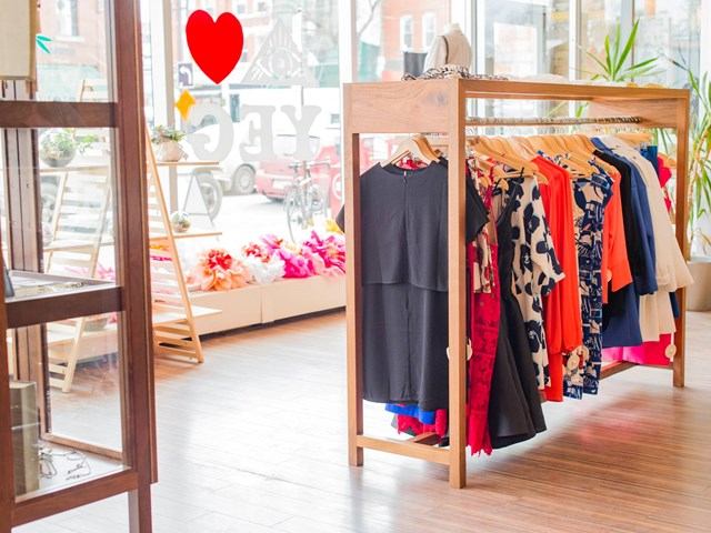 Clothing rack with shirts in a shop window, near storefront displays and potted plants.