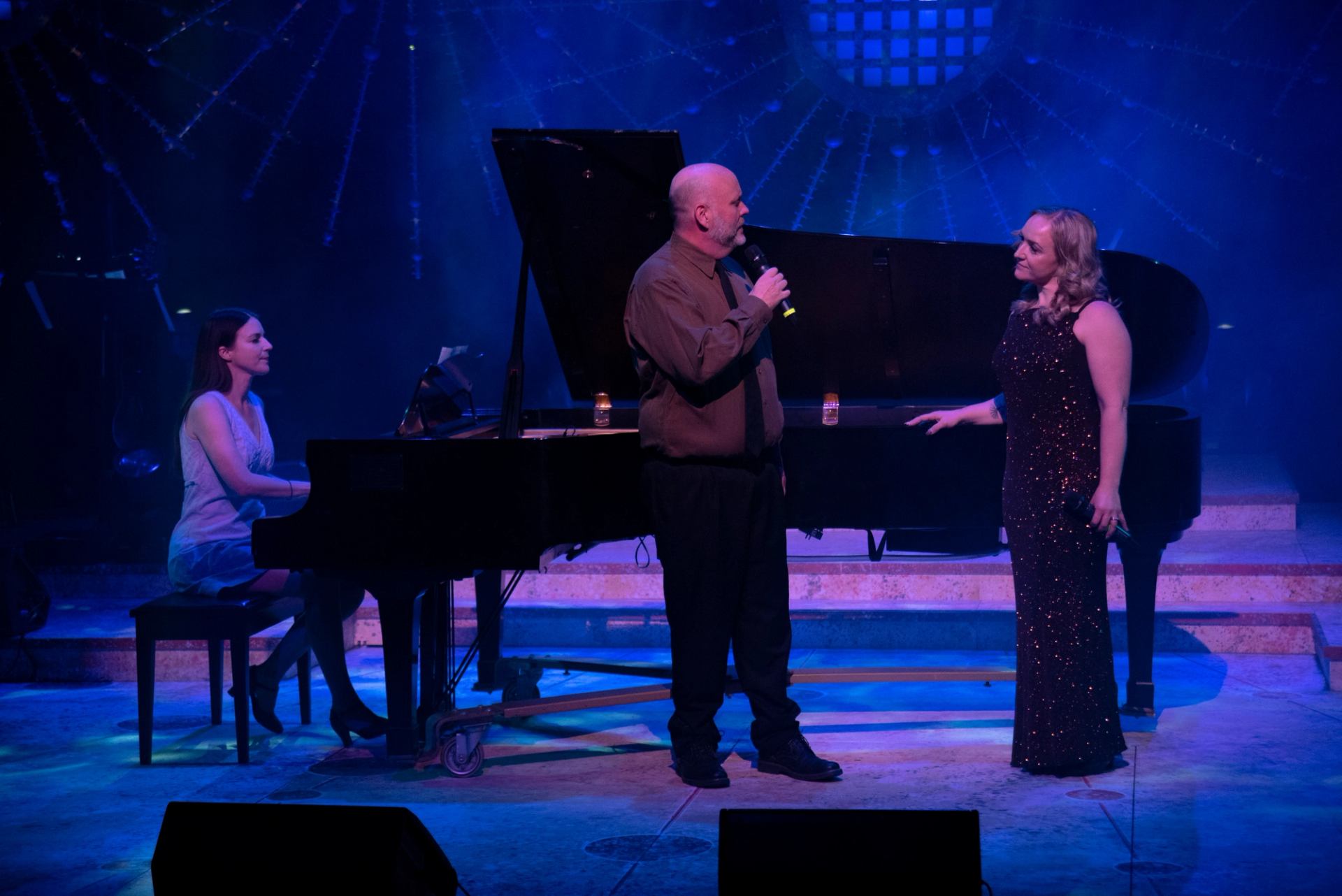 Three performers on stage with piano, microphones, and stage lighting.