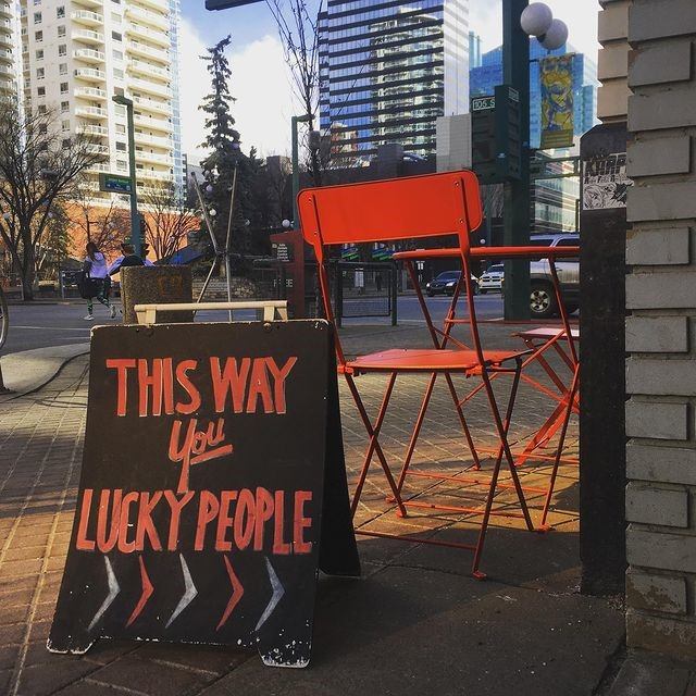 Sidewalk sign reading “This way you lucky people” beside bright orange café chairs.