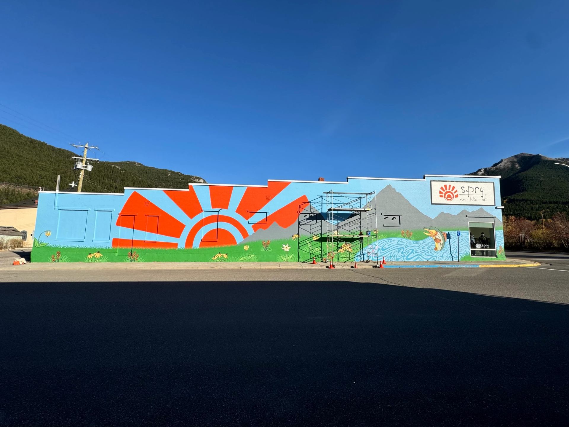 Colourful building mural with sunrise, mountains, river and fish, scaffolding in front.