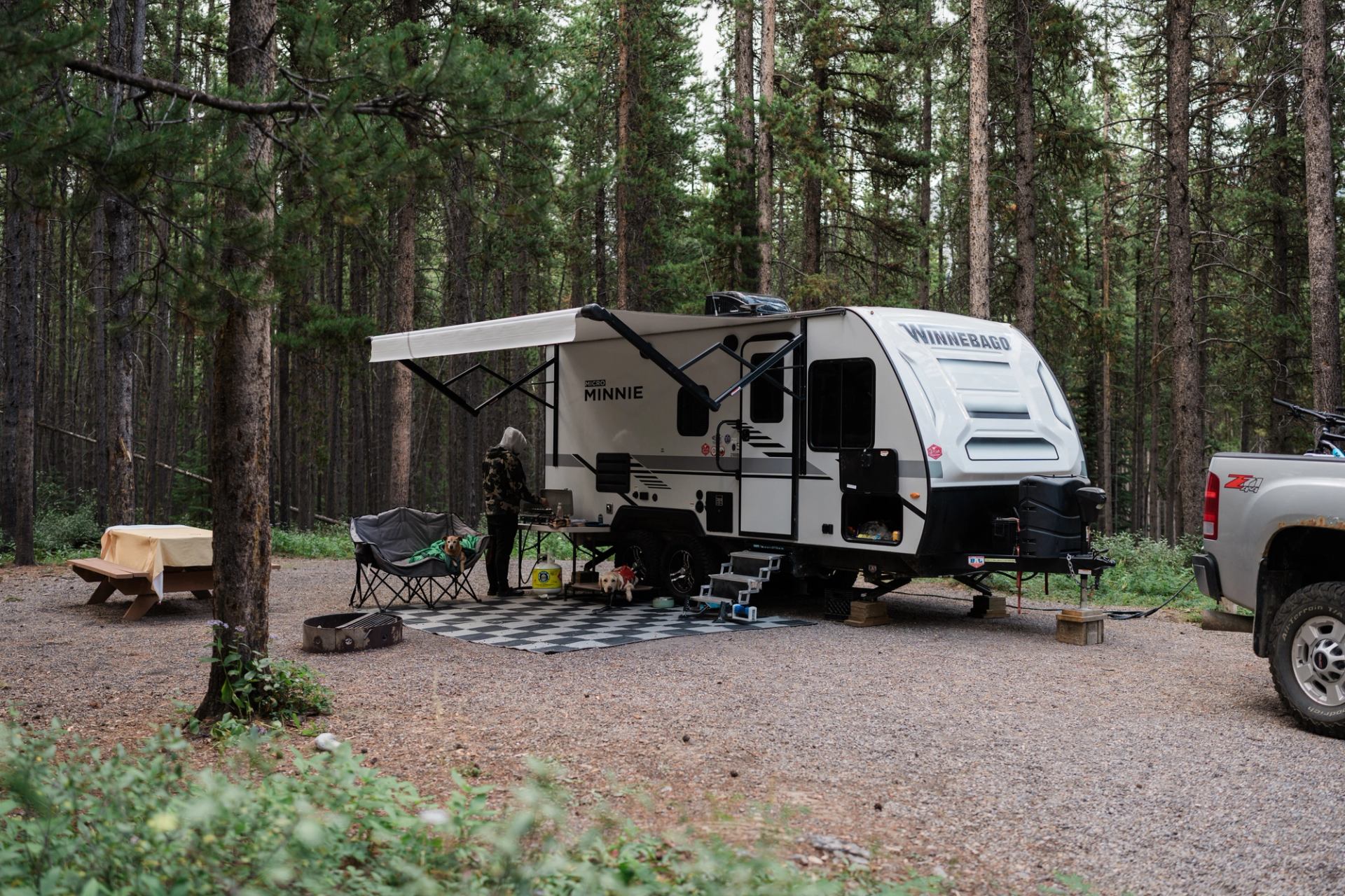 Travel trailer and pickup truck parked at forest campsite with chairs, table and awning.
