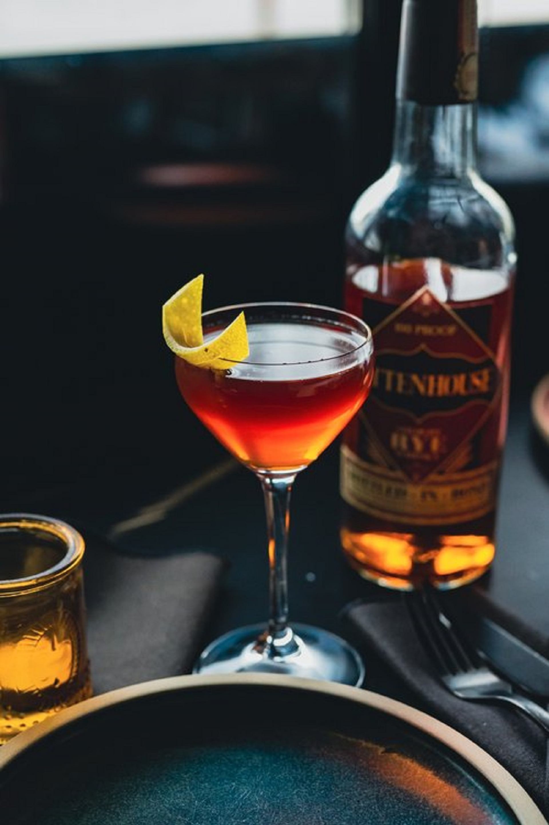 A Manhattan cocktail in a glass with a lemon garnish and a bottle of whiskey behind it.