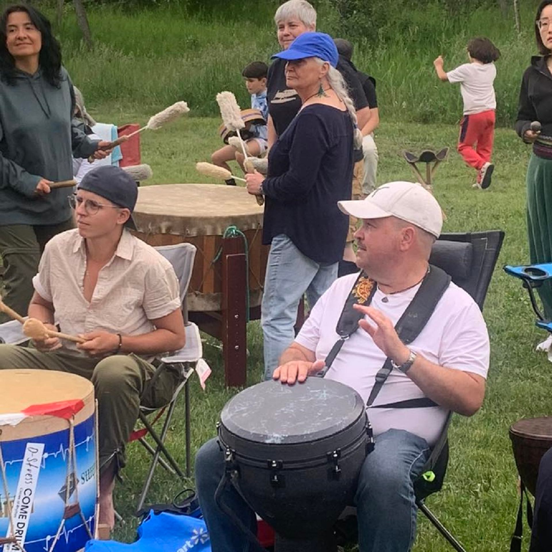 Group of people seated outdoors playing hand drums together on grass during a Circles of Rhythm gathering.