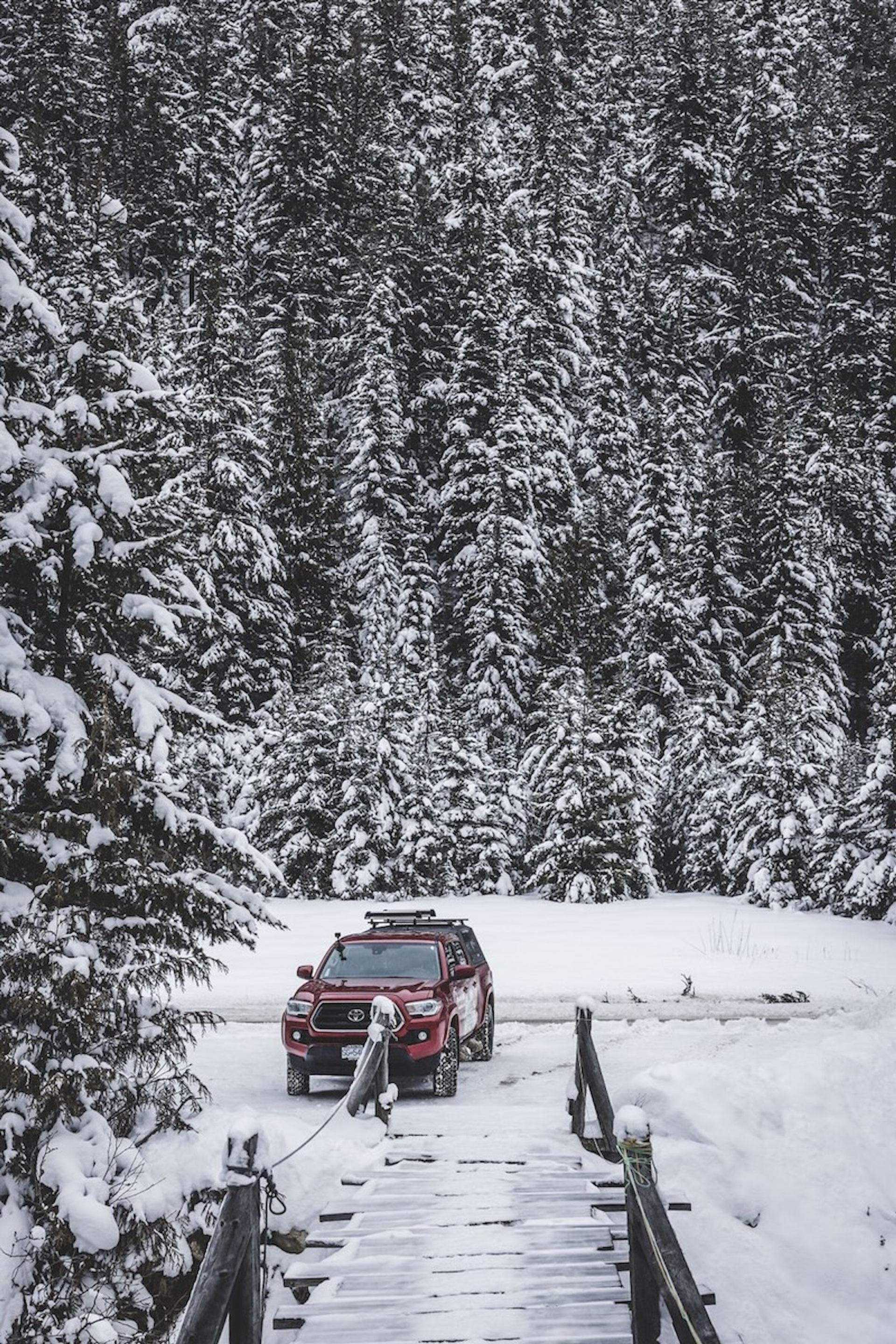 Red 4x4 vehicle crossing a snowy wooden bridge with snow‑covered trees in the background.
