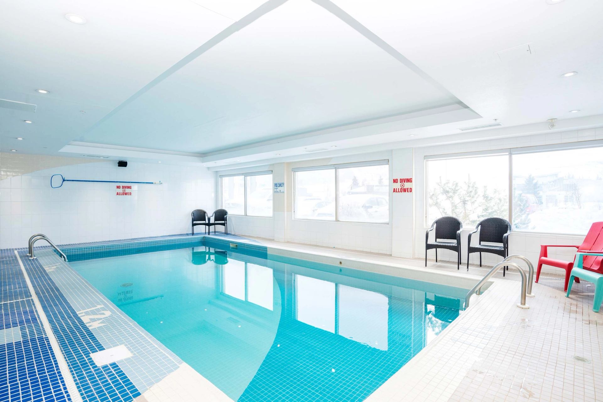 Indoor swimming pool with blue tiles, pool ladder, chairs and large bright windows.