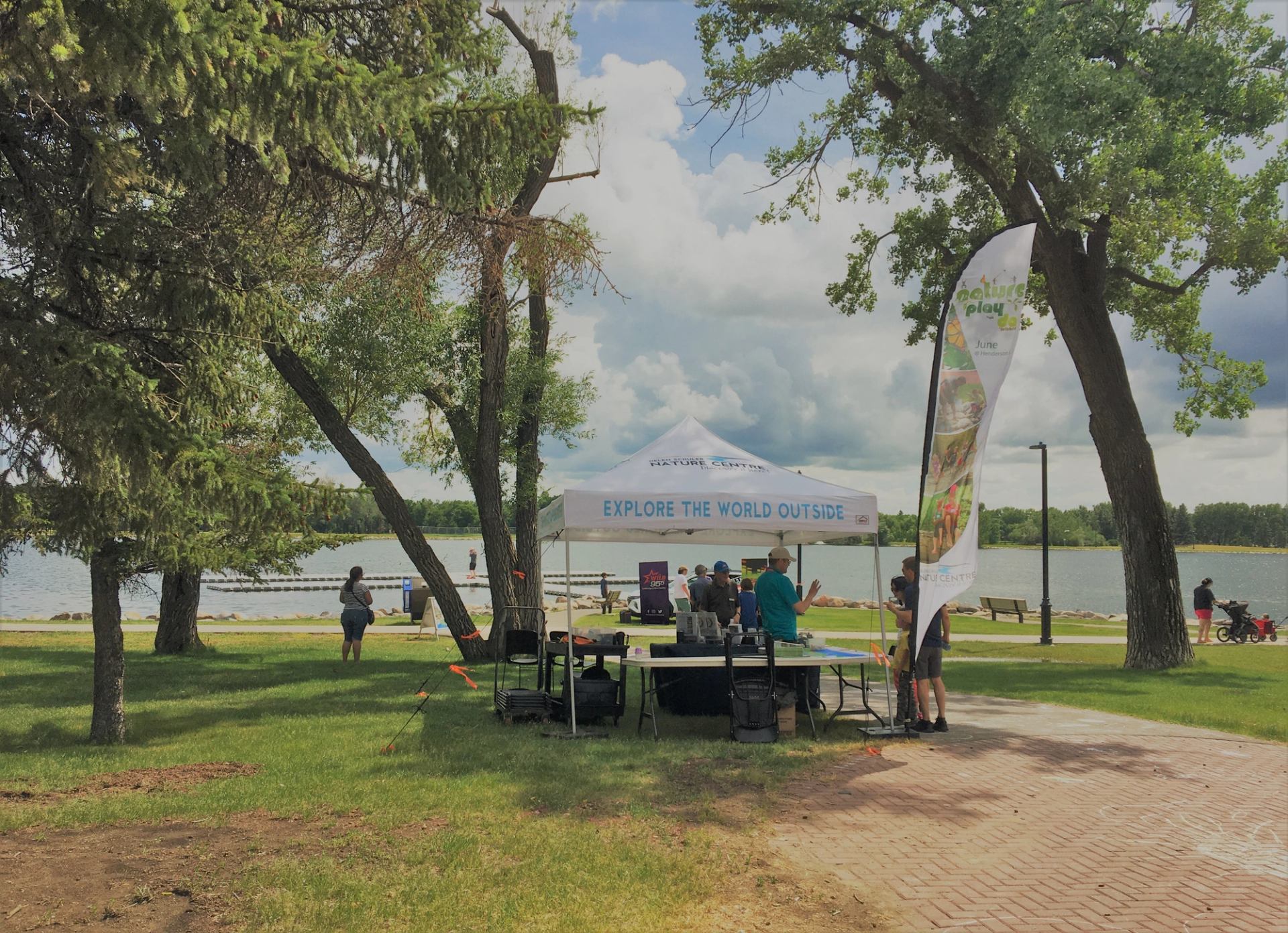 Outdoor event tent reading “Explore the World Outside” beside lakeside path and trees.