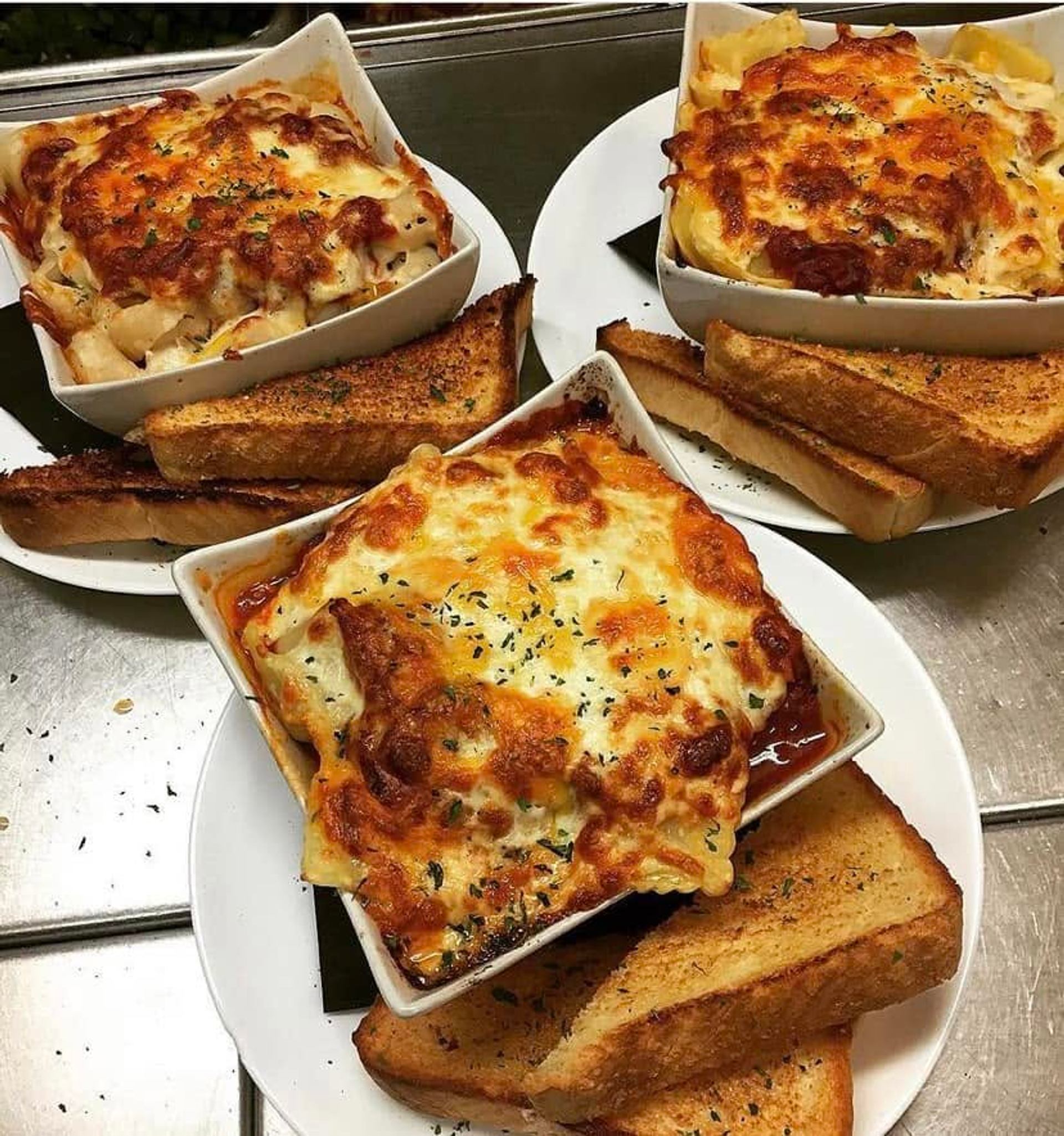Three baked pasta dishes topped with melted cheese are served with slices of garlic toast.