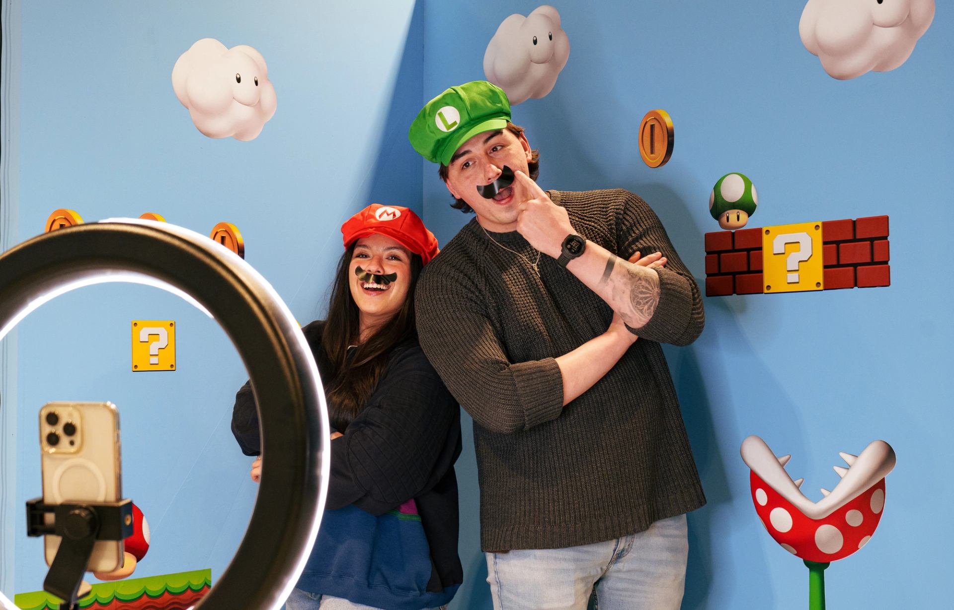 Two people in Mario costumes pose before smartphone, with Super Mario wall decorations.
