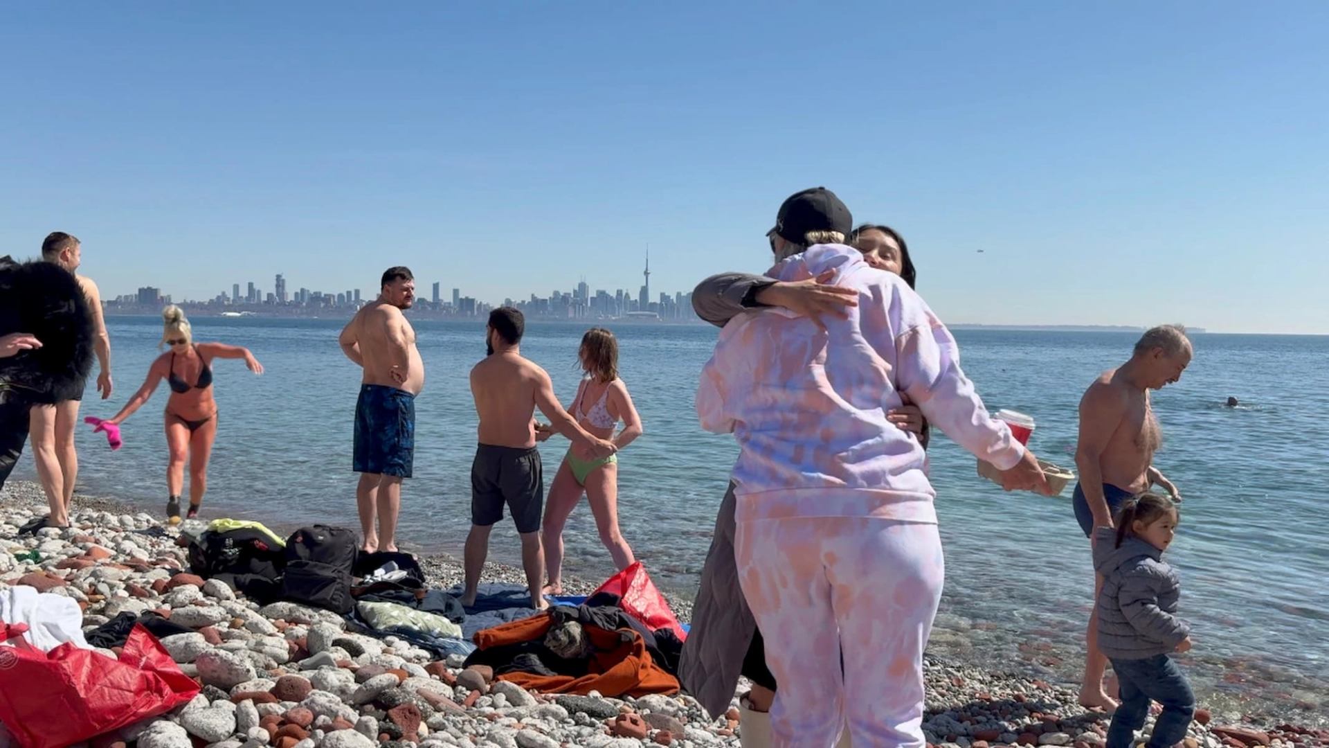 People gather on a rocky lakeshore with Toronto skyline visible across the water.