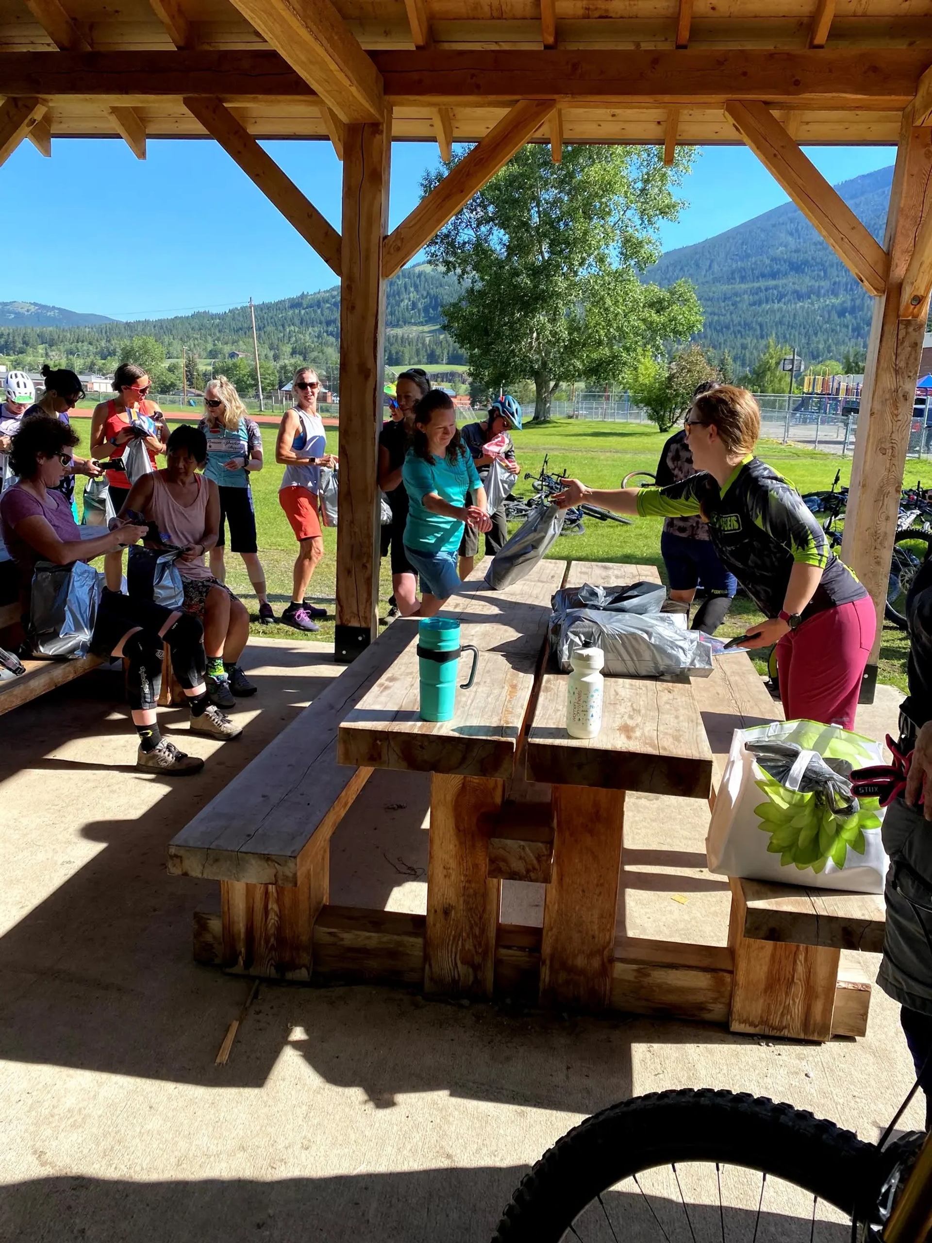 Cyclists gather under a wooden shelter, talking around picnic tables with gear bags