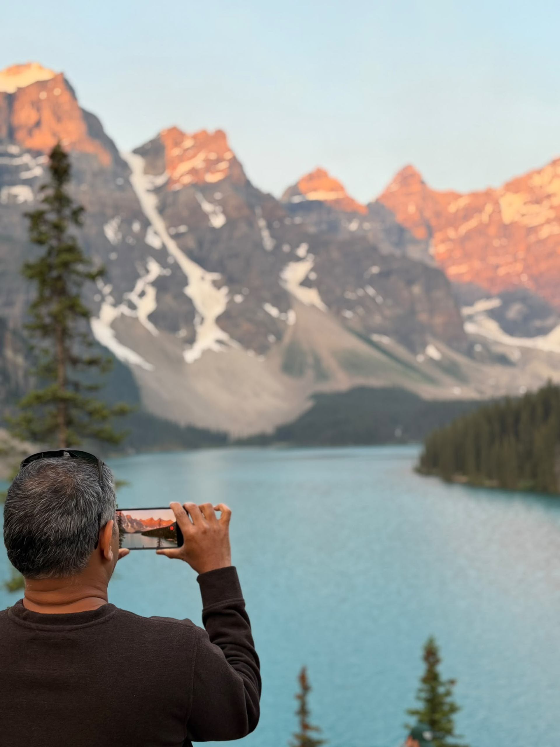 Person photographs a turquoise lake and snow-streaked mountains with a smartphone.