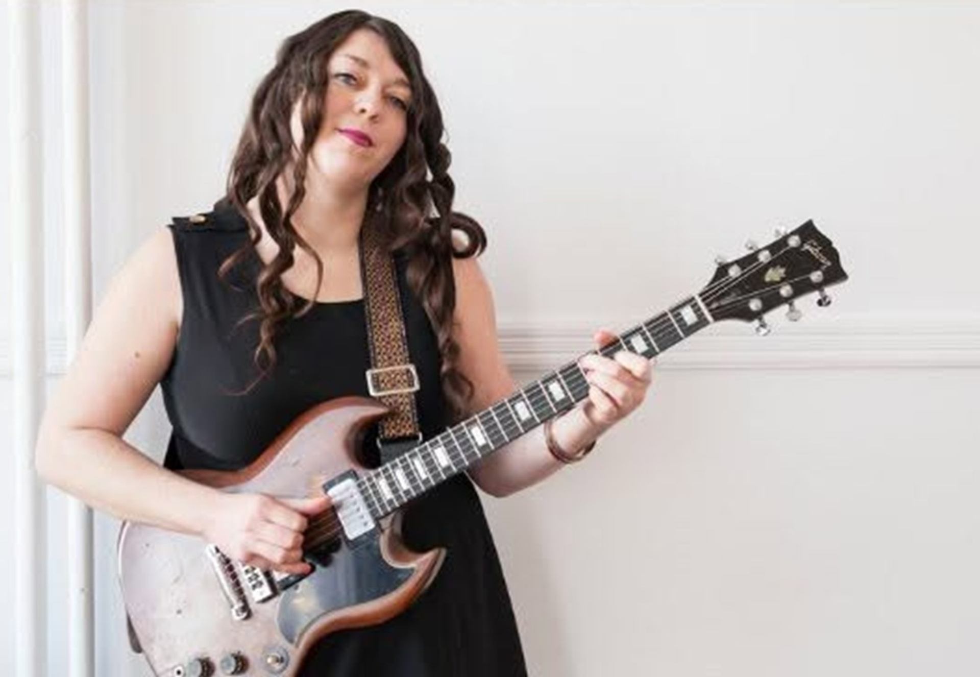 Person in black sleeveless dress playing an electric guitar against a plain white wall.