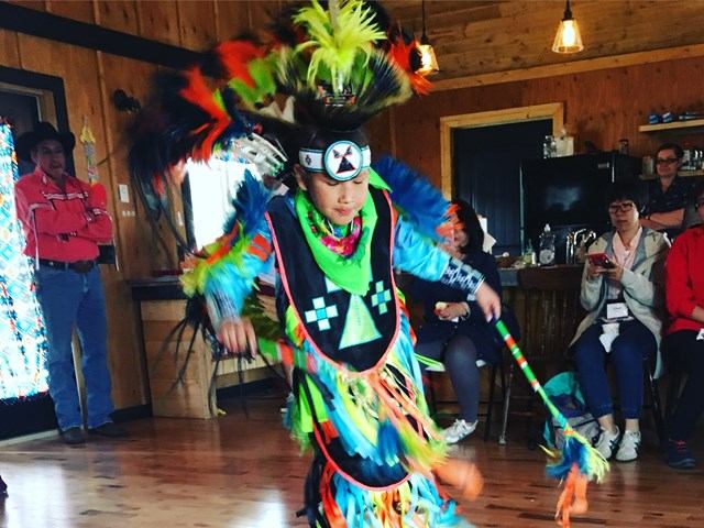 A person performs a traditional Native American dance indoors, wearing vibrant regalia with feathers and beadwork, exuding energy and cultural pride.