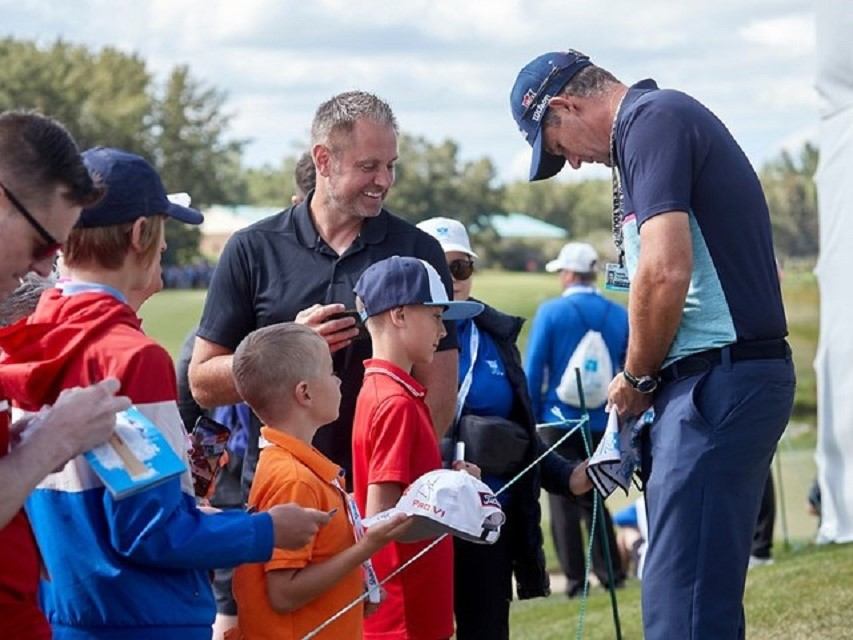 Golfer signs autographs for children holding caps beside a rope at a golf course.