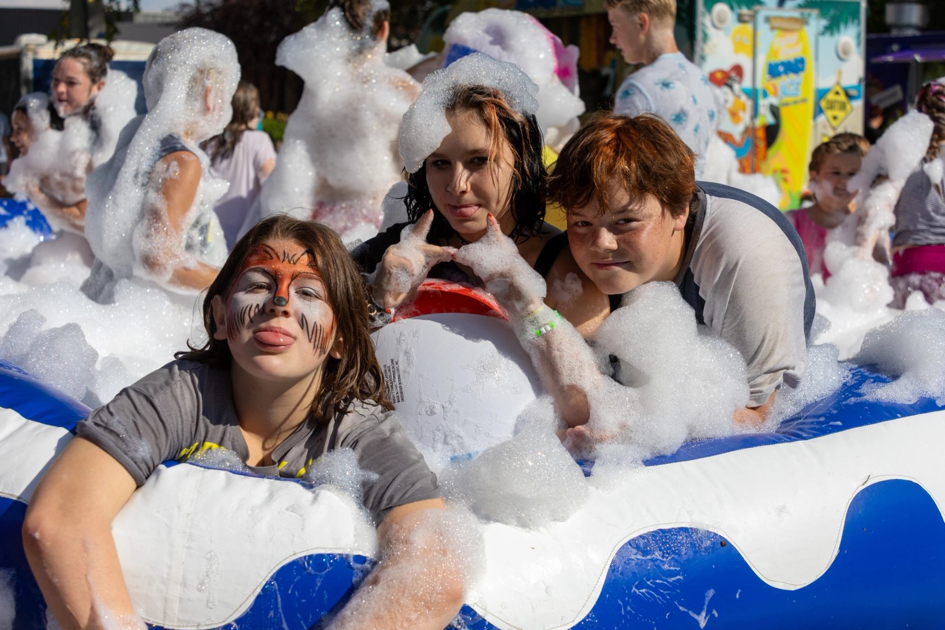 Children with face paint play in foam-filled inflatable pool at outdoor event.