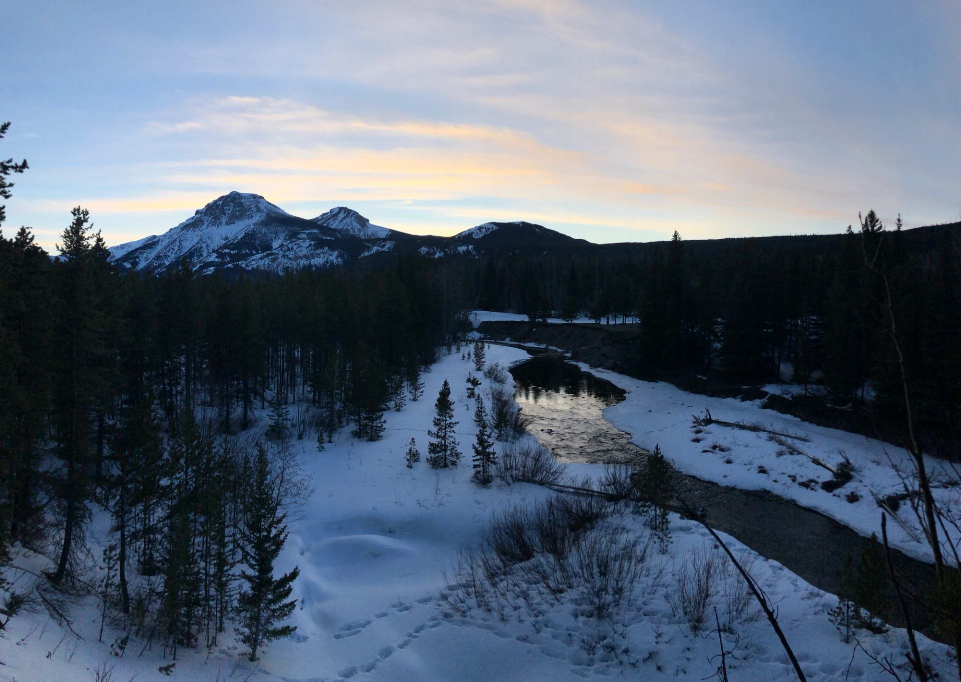 Snow-covered river winding through a forest with mountains silhouetted at sunset.