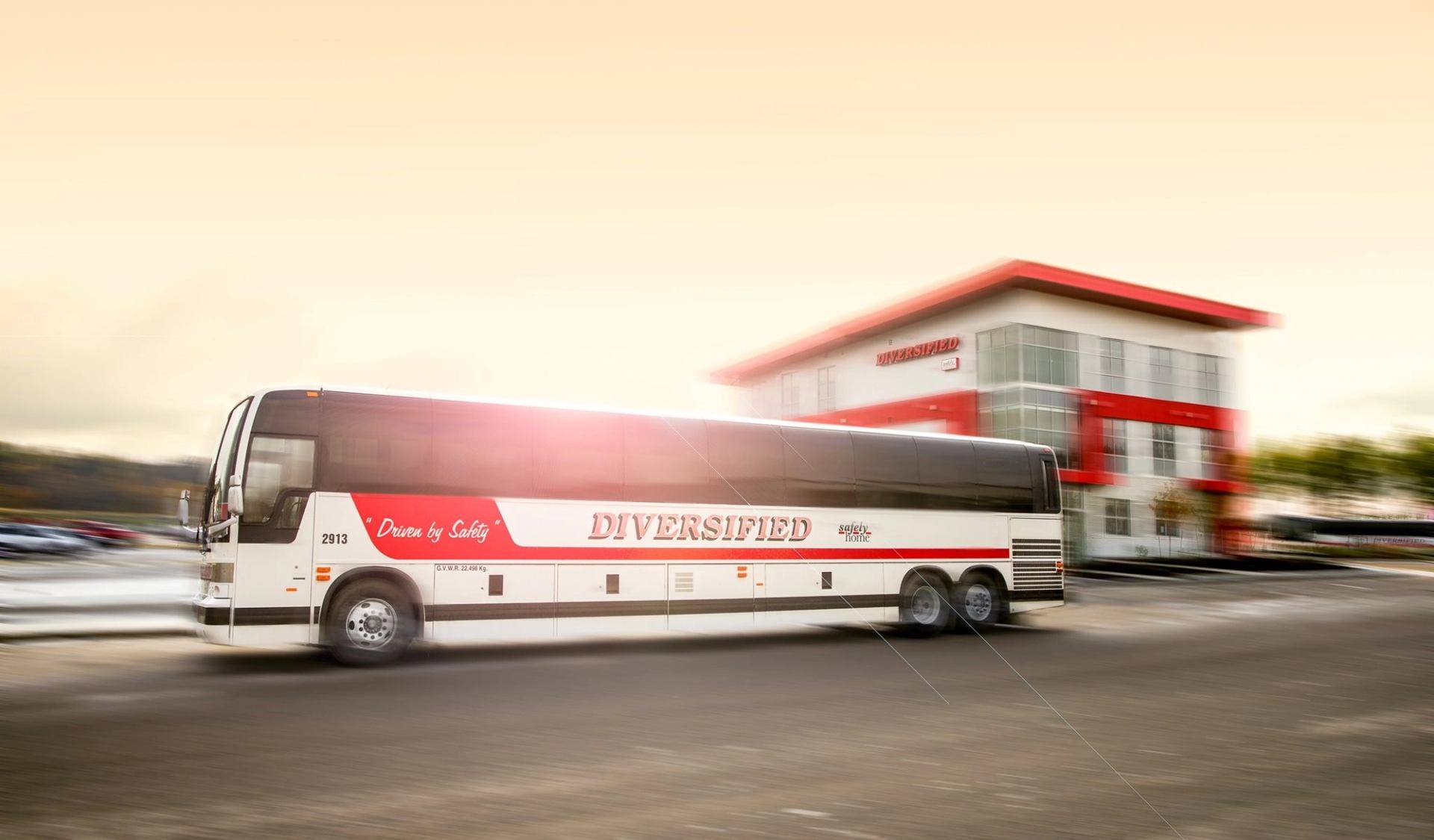 White Diversified bus with red accents and “Driven by Safety” text moves past a modern building.