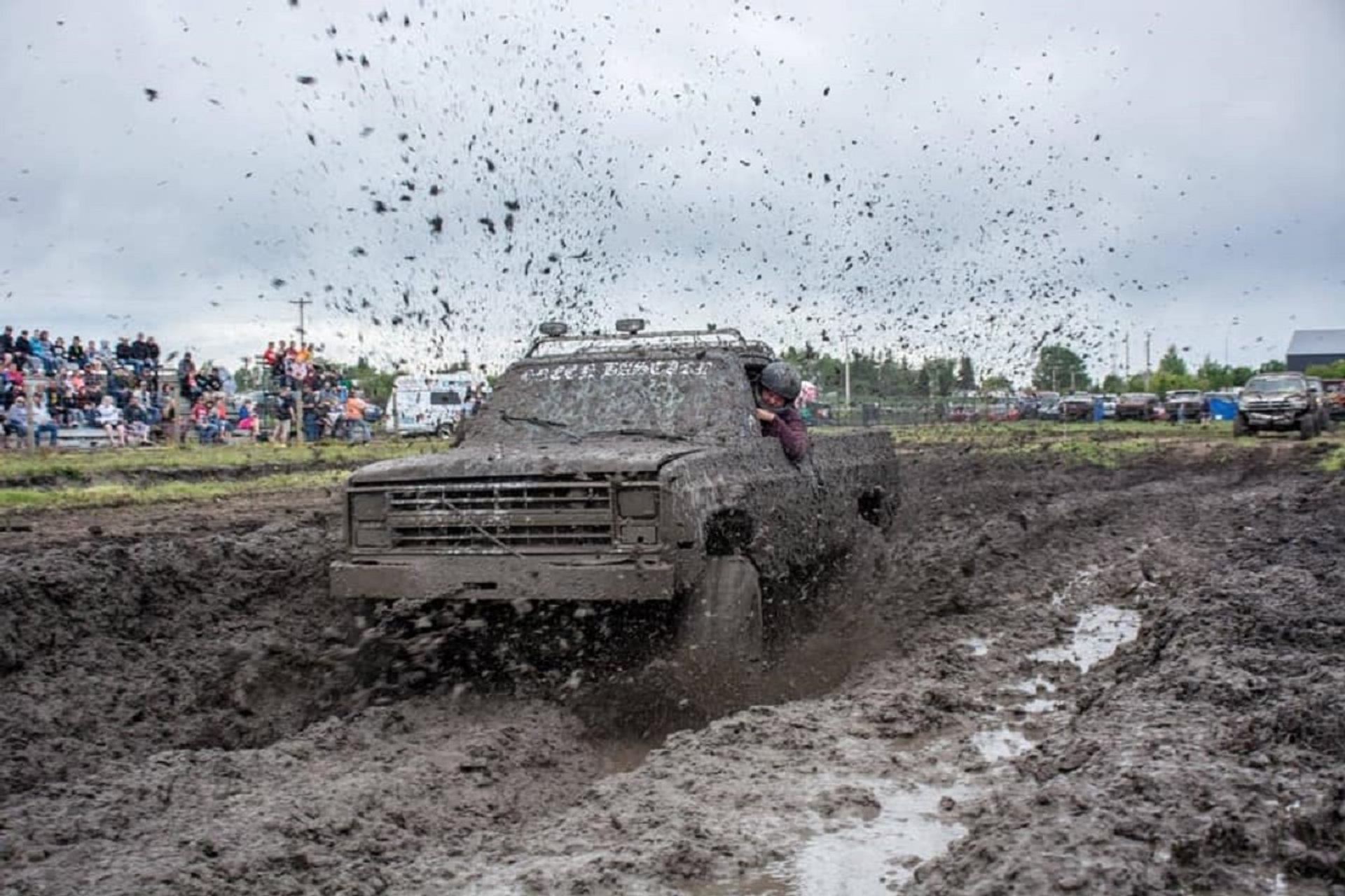 Mud-covered pickup truck drives through deep mud pit as spectators watch from bleachers.