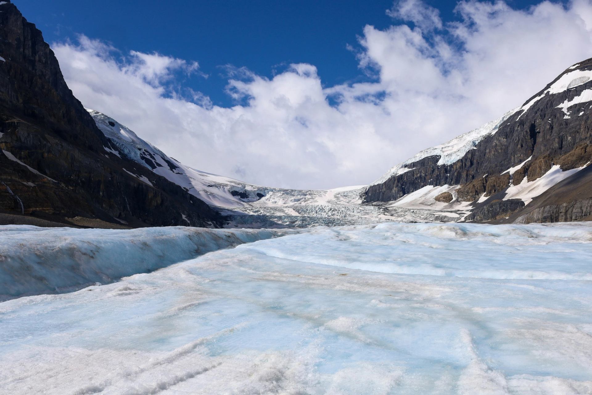 Wide glacier between rocky snow-covered mountains under a bright blue sky with clouds.