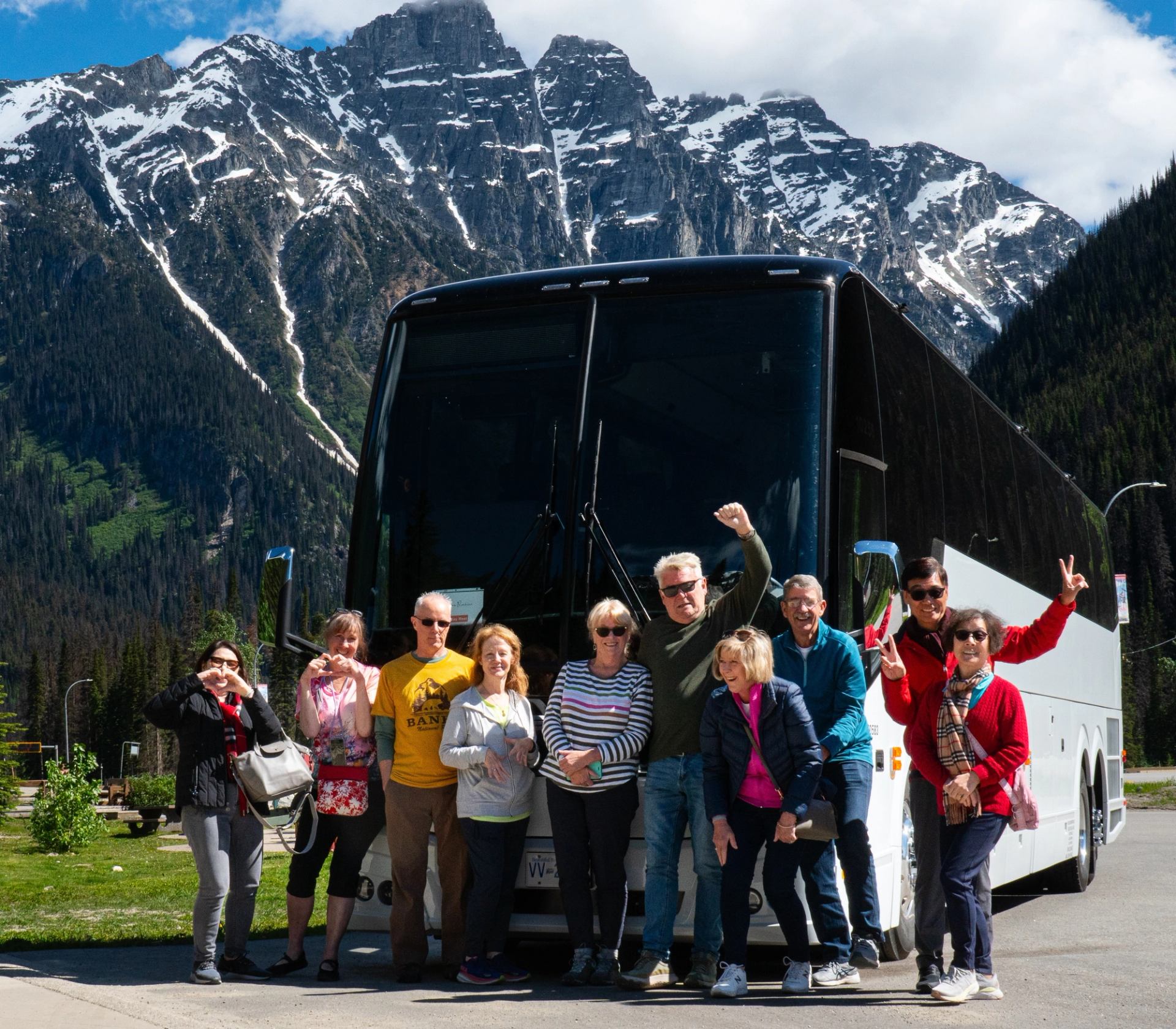 A group standing in front of a large tour bus with tall snow‑capped mountains behind them.