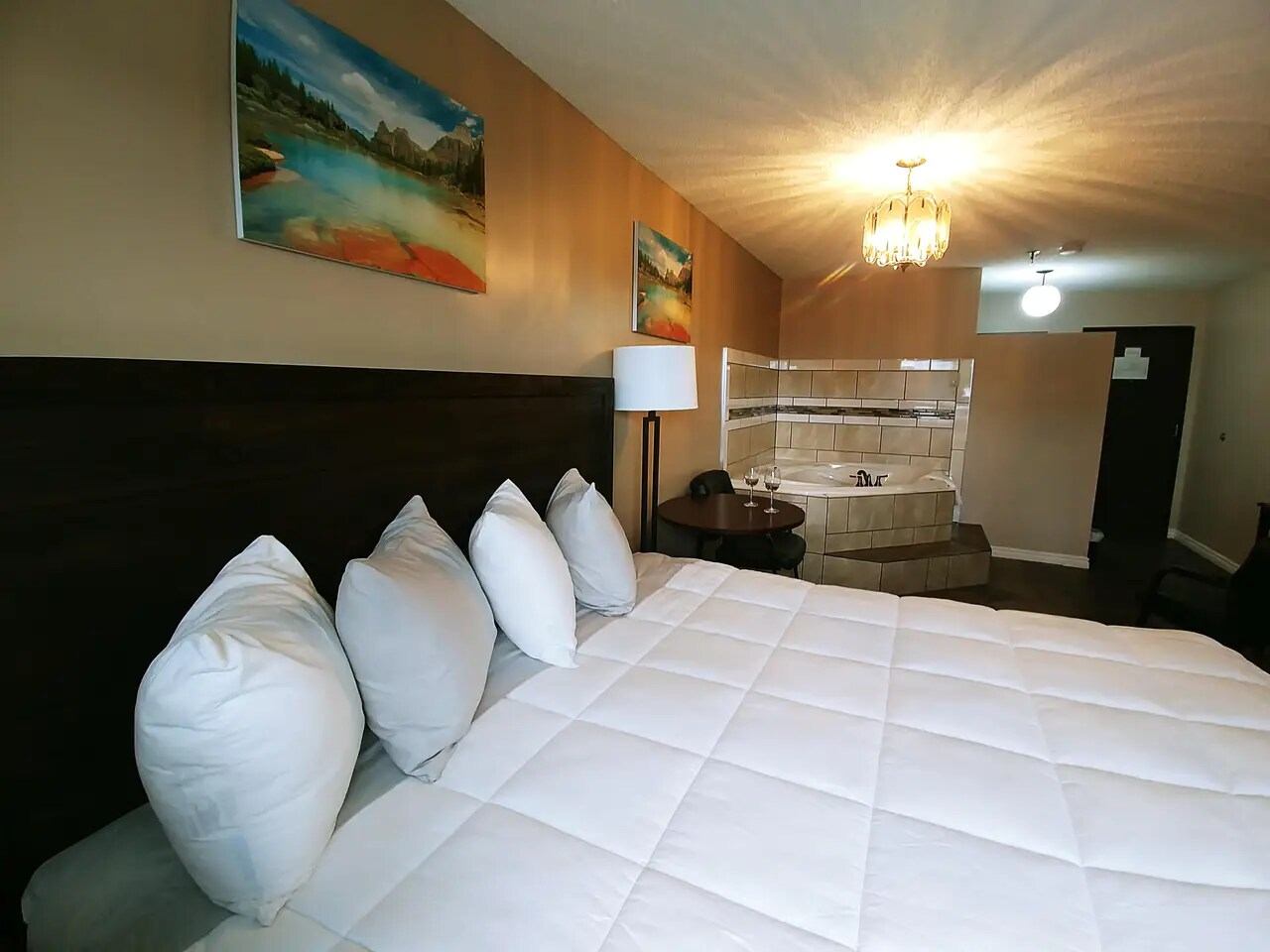 Spacious guest room featuring a plush bed, warm lighting, artwork, and a tiled in-room jacuzzi.