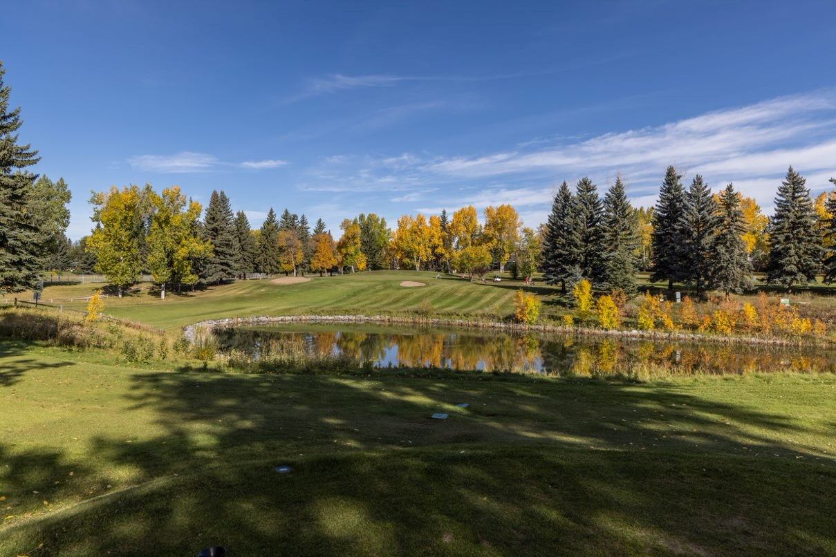 Golf course fairway beside pond, sand bunkers, and yellow-leaved trees under blue sky.