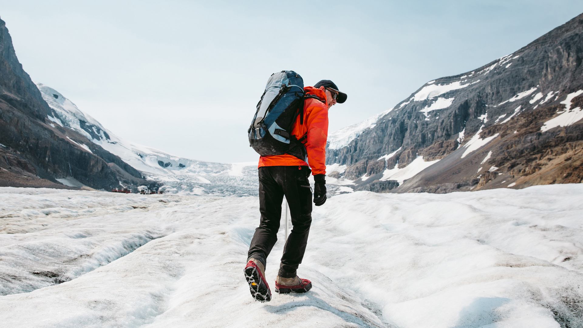 Hiker in orange jacket stands on glacier with boots, trekking pole, and mountains in background.