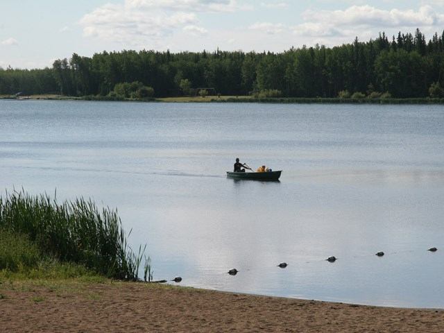 Two people row a small boat across the still lake near the sandy shoreline.