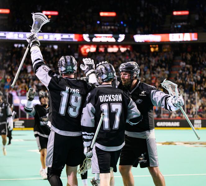 Lacrosse players in black uniforms celebrate on indoor field with crowd and scoreboard in background.