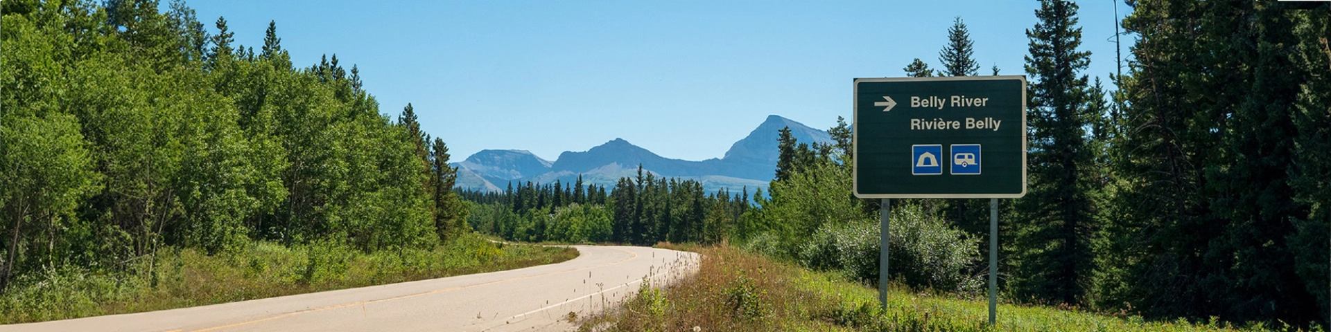 A road sign for Belly River with tunnel and camping icons, next to a road surrounded by trees and mountains.