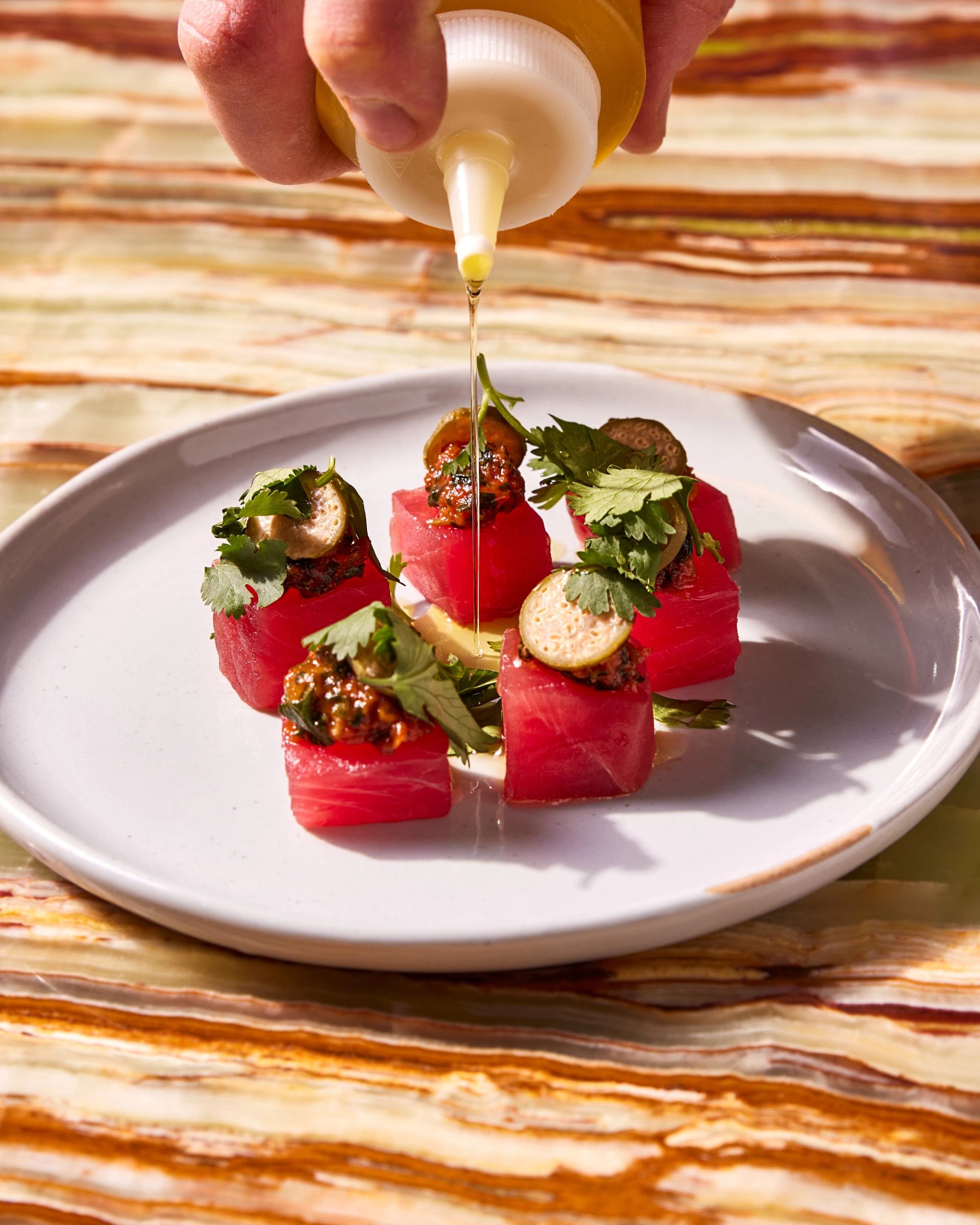 Hand pours sauce from a spoon onto plated red tuna and herbs.