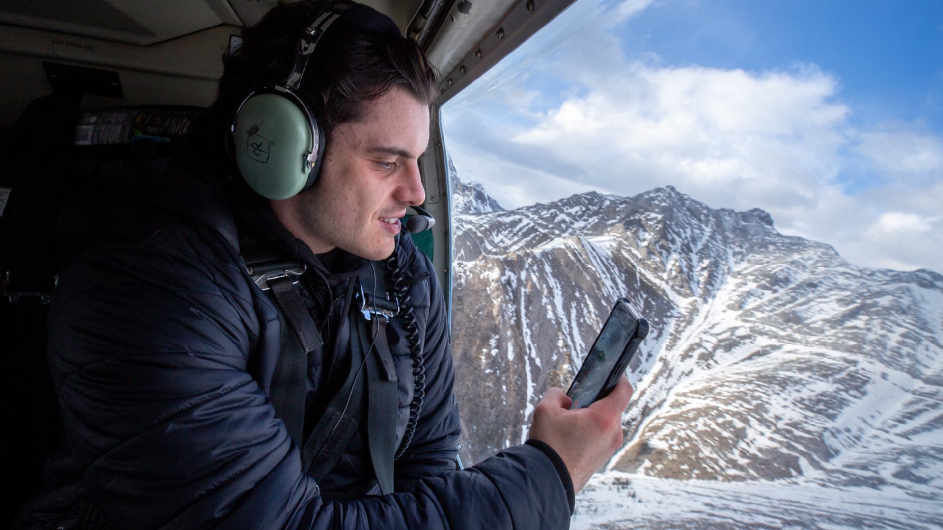 A passenger inside a helicopter looks out at rugged snowy mountain terrain.