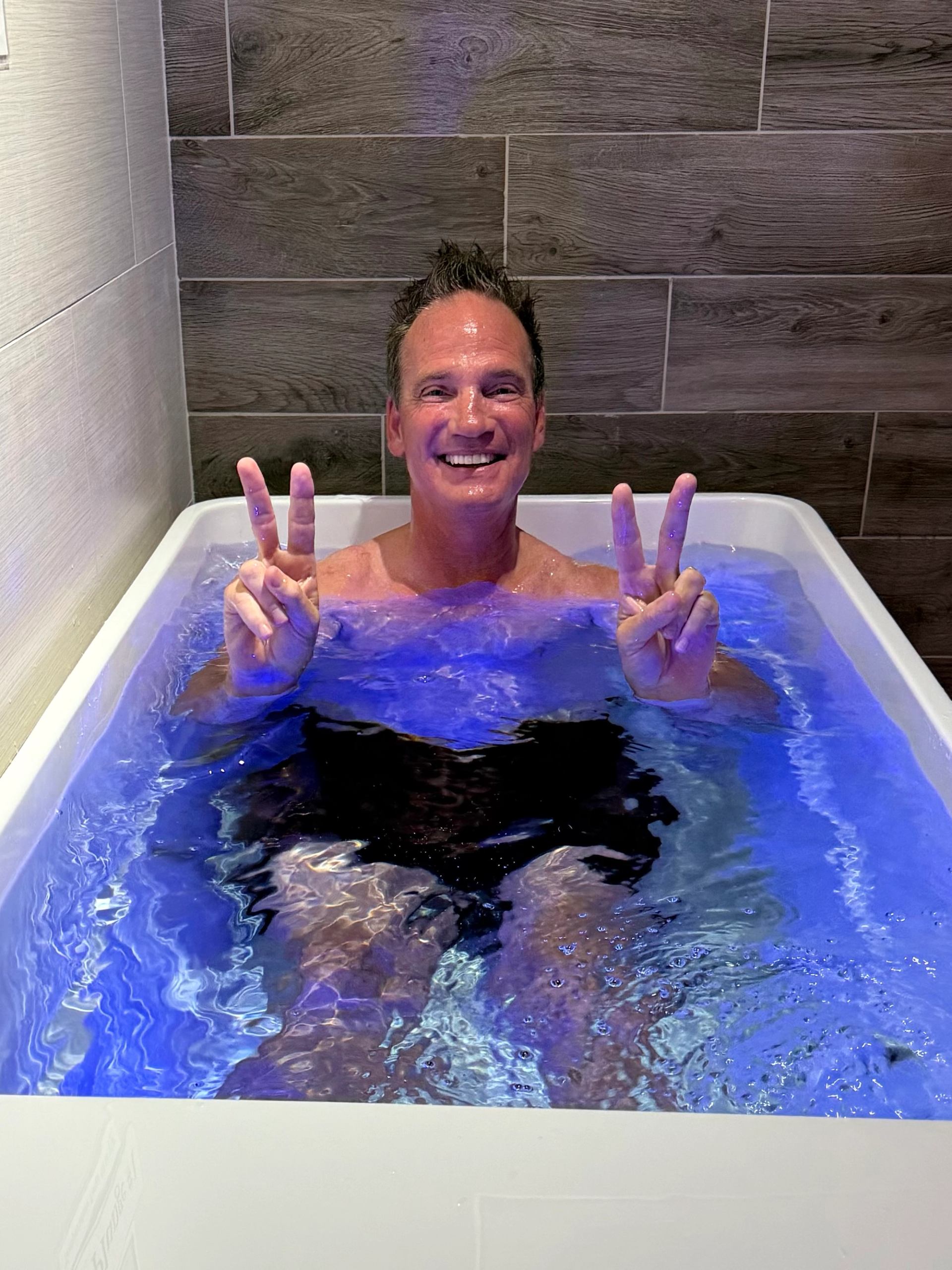 Person in a glowing blue-lit tub, smiling and flashing peace signs in a tiled room.