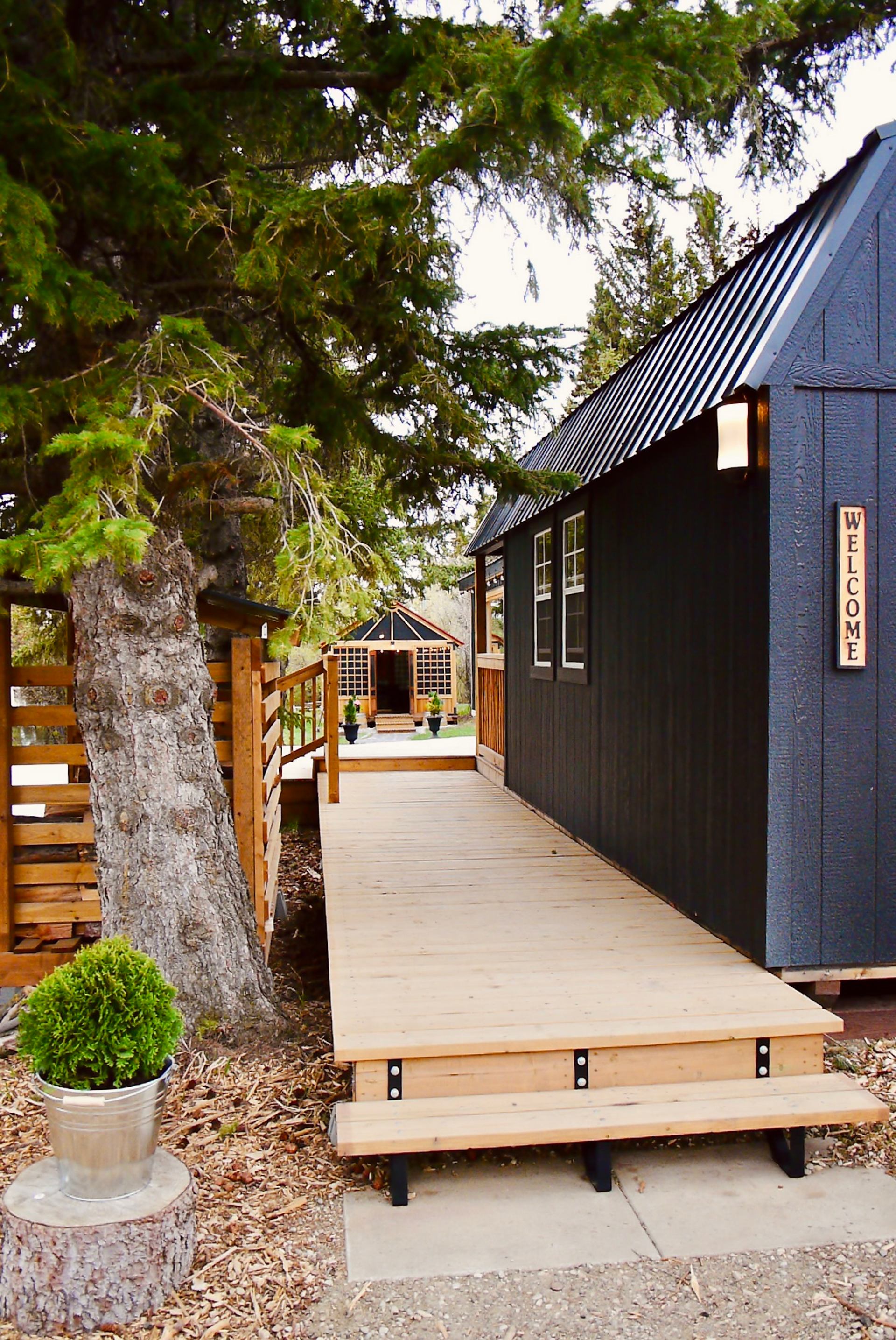A wooden walkway leads past a dark exterior building with a “Welcome” sign toward a sauna structure in the background. Trees and landscaping frame the calm outdoor space.