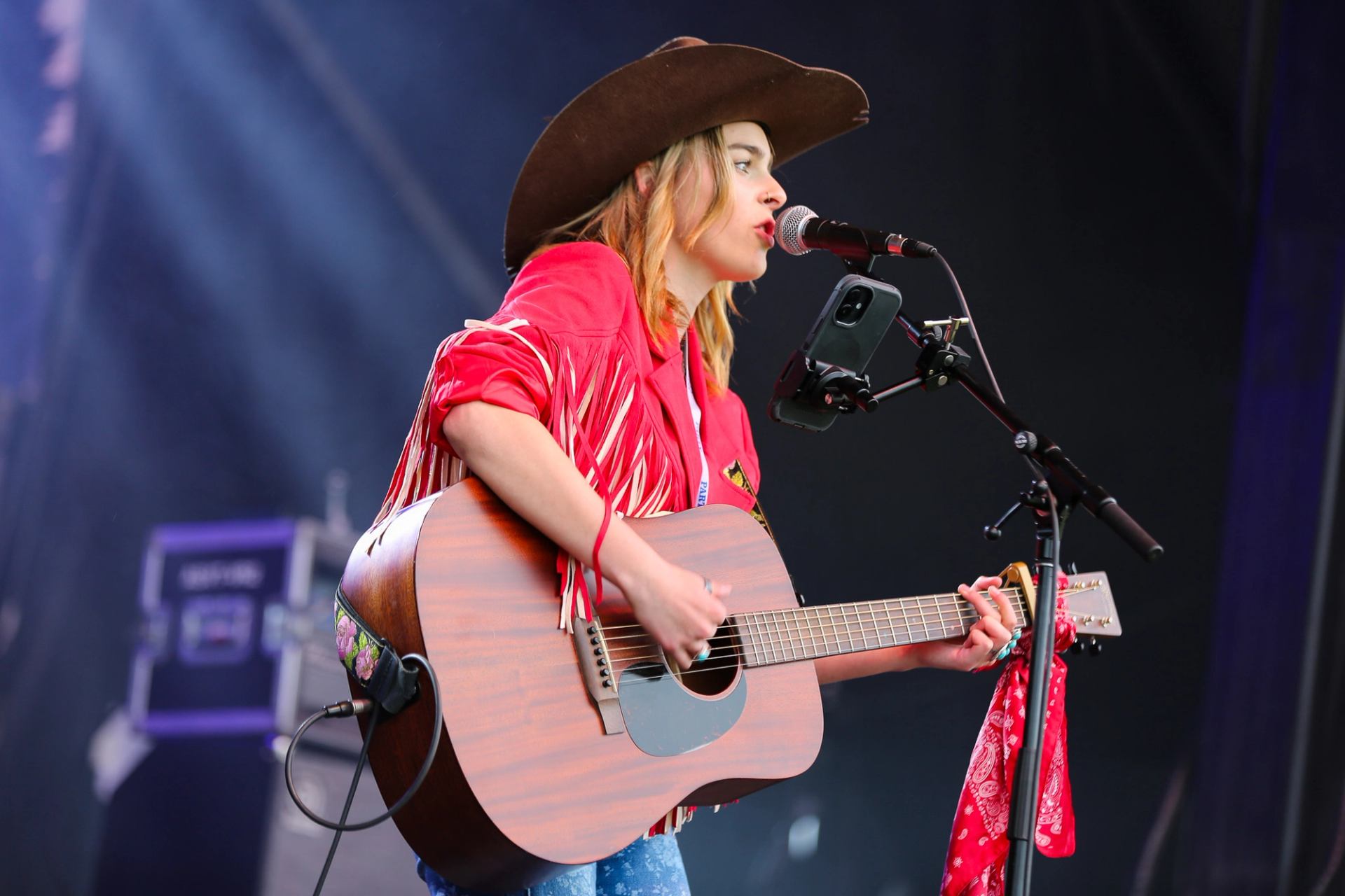 Performer in a cowboy hat playing acoustic guitar onstage with lively red fringe attire.