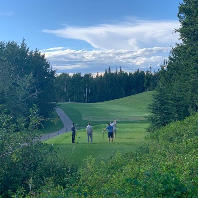 Golfers teeing off at The Dunes Golf & Winter Club with rolling fairways, trees, and a cloudy sky.
