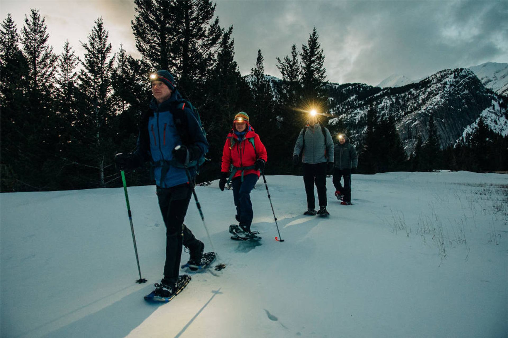 Group walks on snowy trail at dusk with headlamps lighting the path.