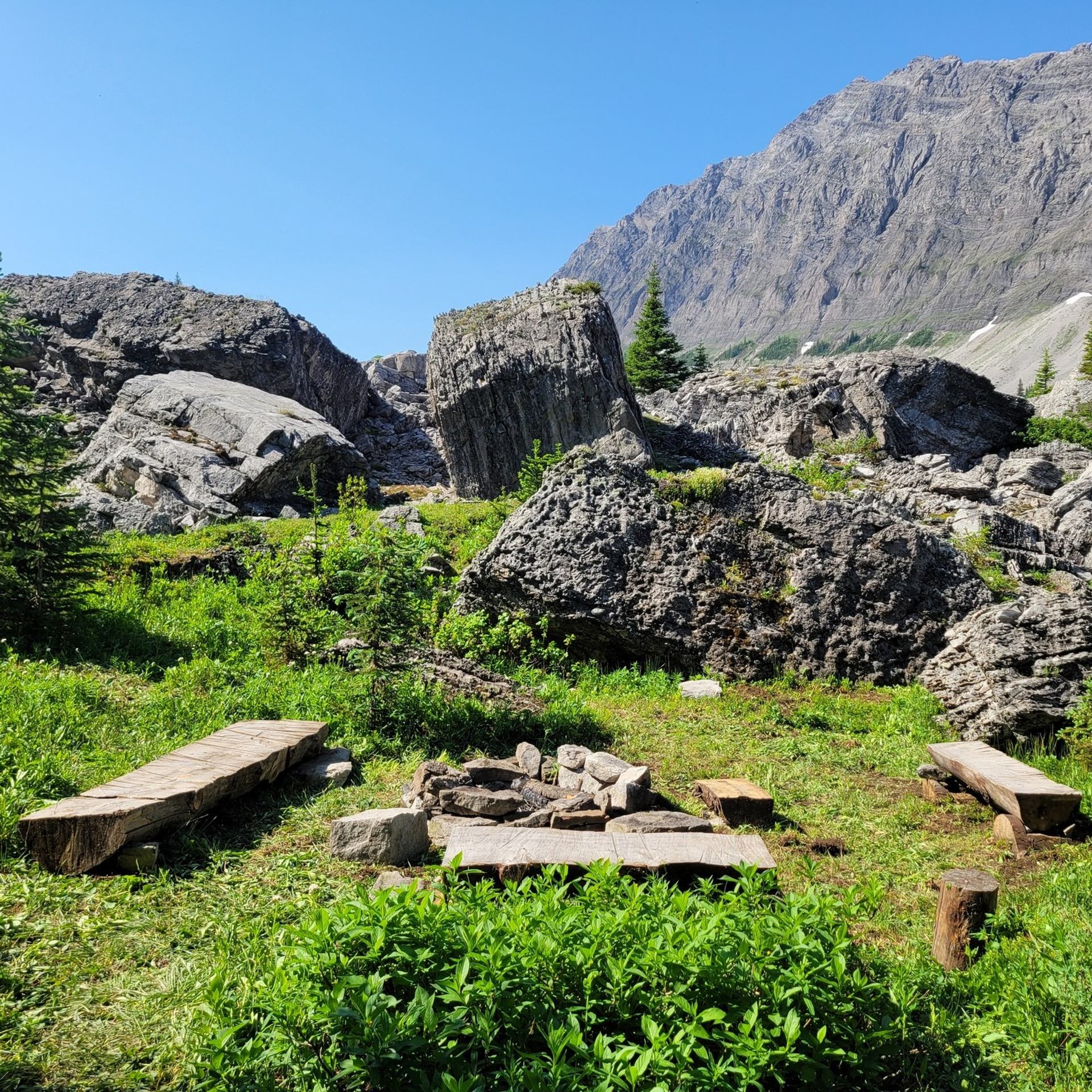 Rocky mountain campsite with wooden benches, fire ring, green grass and clear sky.