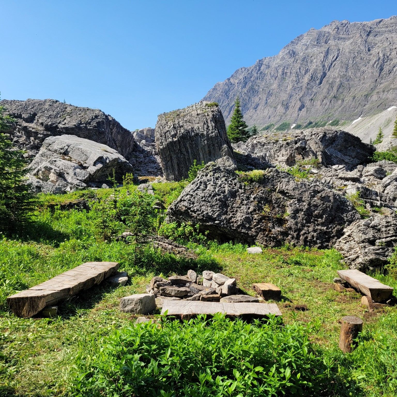 Rocky mountain campsite with wooden benches, fire ring, green grass and clear sky.