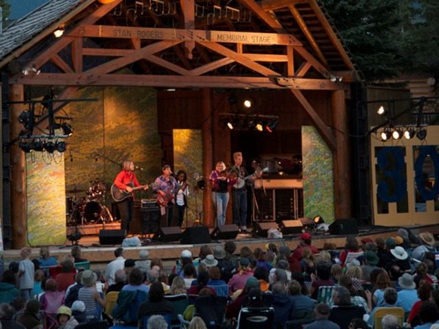 Outdoor band performs on wooden memorial stage before seated crowd at music event.